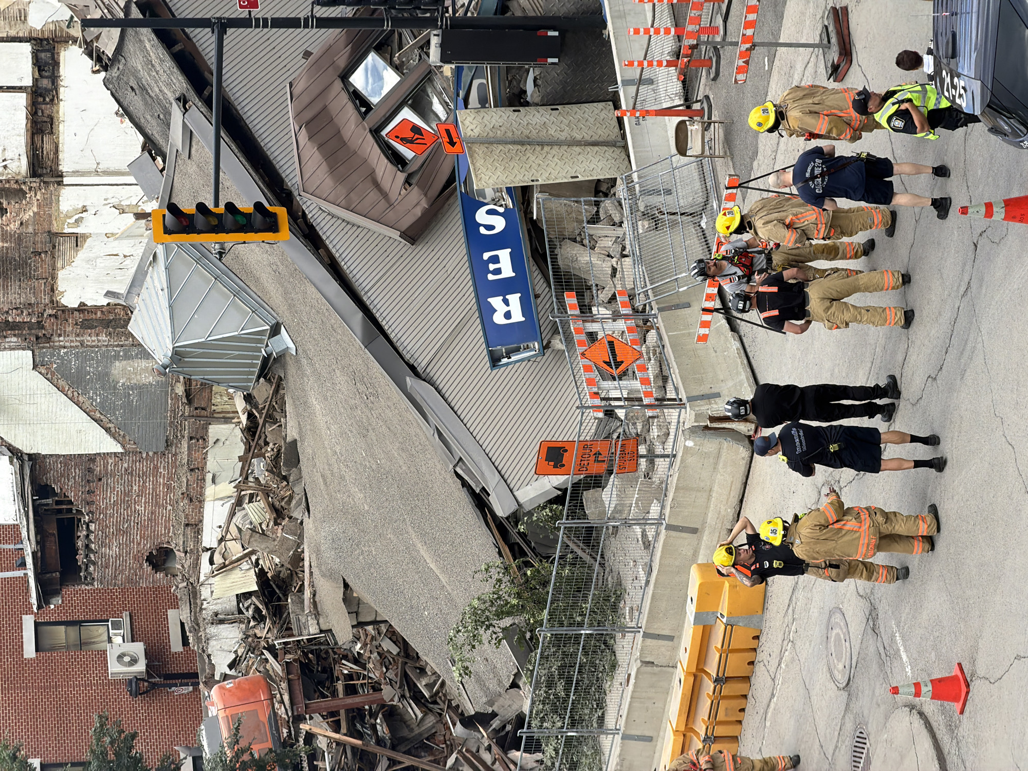 The image shows a scene of a collapsed building, with emergency personnel present. Several firefighters in full gear, wearing yellow helmets and tan turnout suits, are visible. Some are standing, while others appear to be walking or conferring. One firefighter is wearing a black t-shirt and dark shorts, and another is wearing a high-visibility vest. Traffic cones and barricades are set up, indicating a controlled area. A traffic light is visible, with its red, yellow, and green lights illuminated. Several orange construction signs are also present, one reading "DETOUR" with an arrow, and another with a truck icon and an arrow. The building itself appears heavily damaged, with exposed brick and debris scattered around. The sky is overcast, suggesting a cloudy day. The scene suggests an ongoing emergency response to a building collapse, likely in an urban environment. Text on the firefighter's shirt reads "FIRE RESCUE". A license plate on a vehicle reads "21-25".