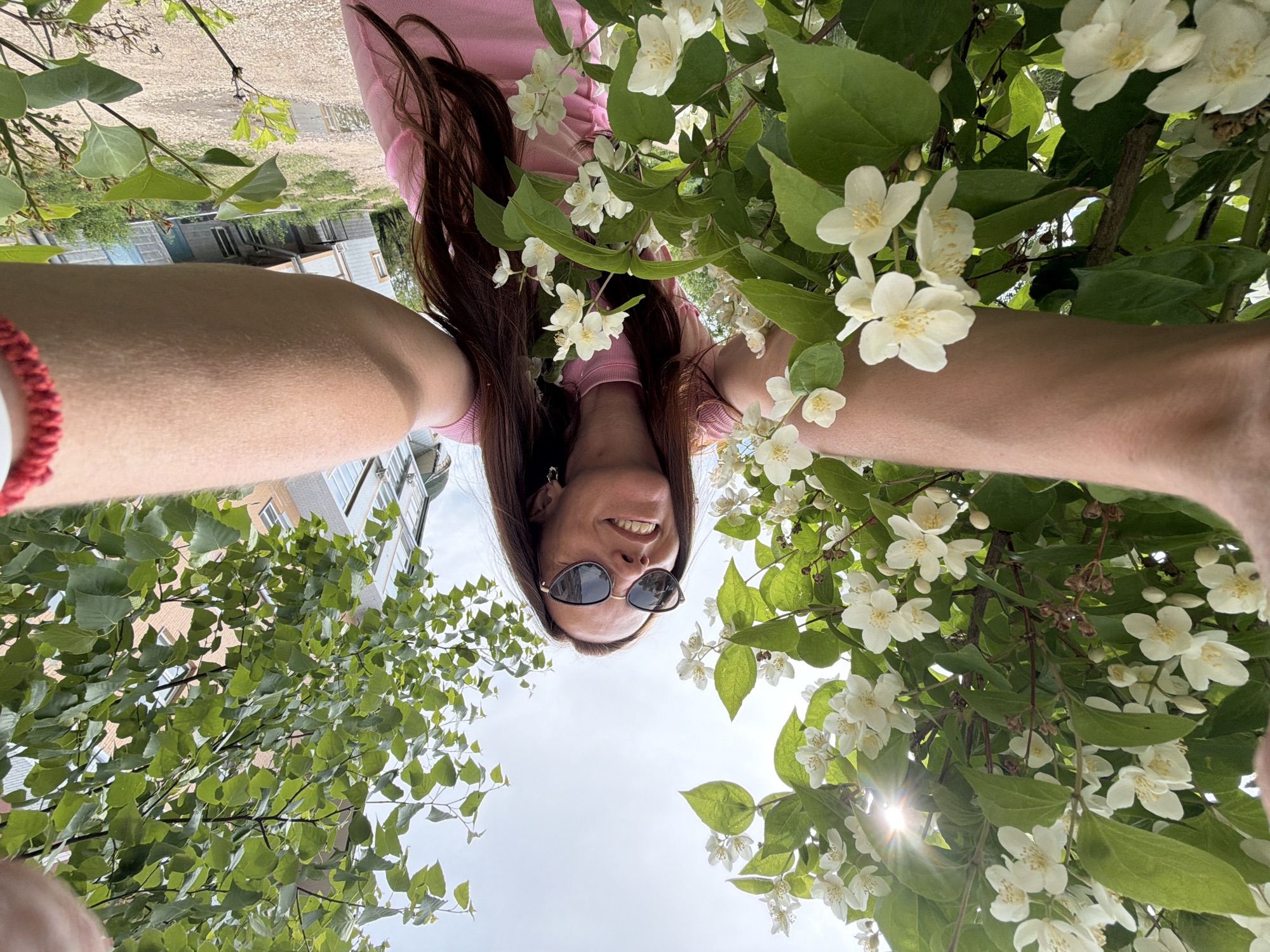 A low-angle, wide-shot photograph captures a woman taking a selfie amidst blooming white jasmine flowers and green foliage. The woman, wearing sunglasses and a pink t-shirt, is smiling and looking upwards. Her arms are extended towards the camera, holding the device. The background shows a building and a cloudy sky. The scene is consistent with an outdoor setting in Birobidzhan, Russia, likely during the warmer months when jasmine blossoms.