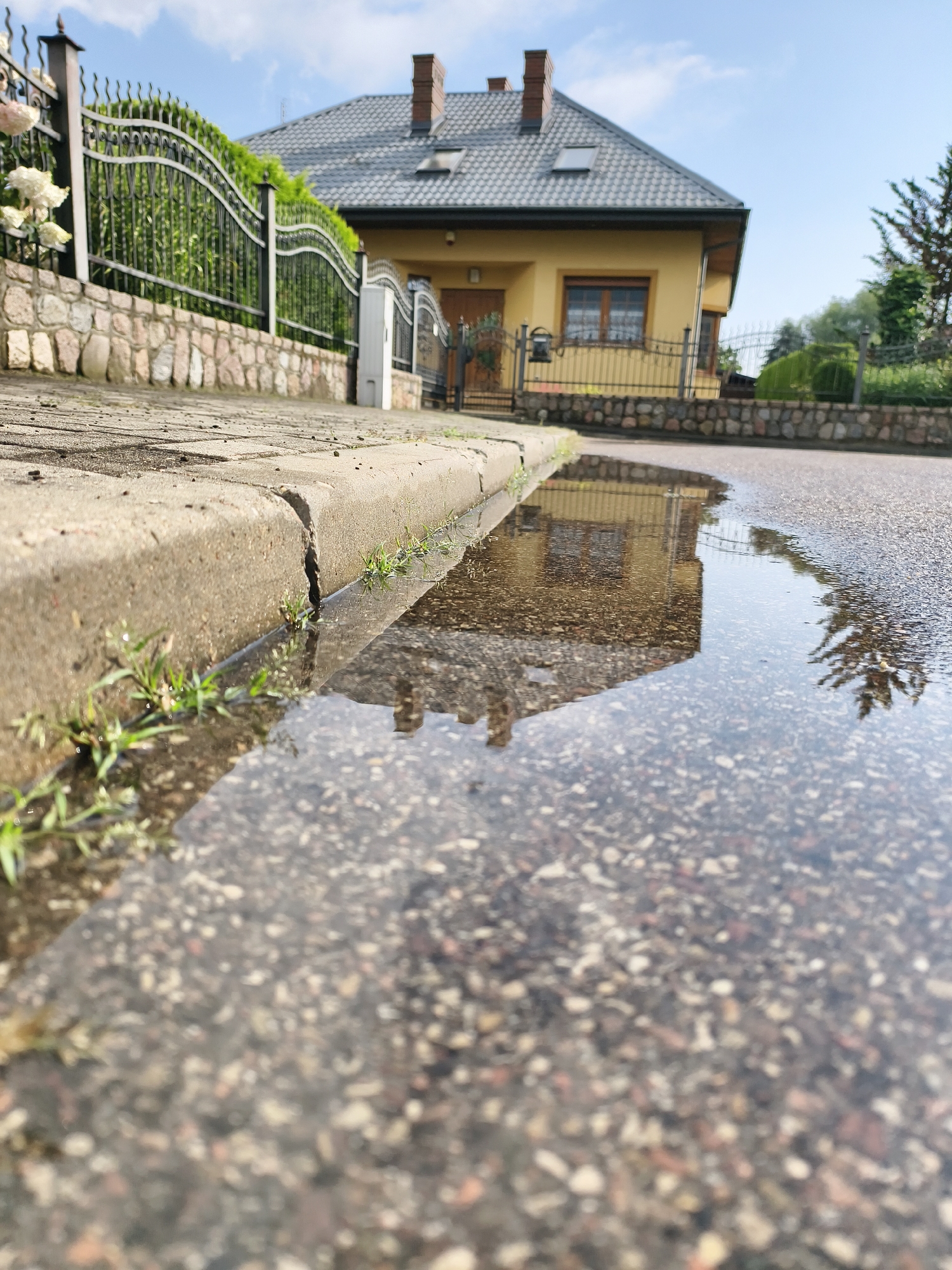 A low-angle shot captures a cracked concrete curb bordering a wet asphalt road in Tczew, Poland. A shallow puddle reflects a yellow house with a dark gray roof and a black metal fence. Small green weeds sprout from the crack in the curb and along its edge. The background features a stone wall, a neatly trimmed green hedge, and a clear sky with scattered clouds. The overall impression is one of a suburban residential street after a rain shower.