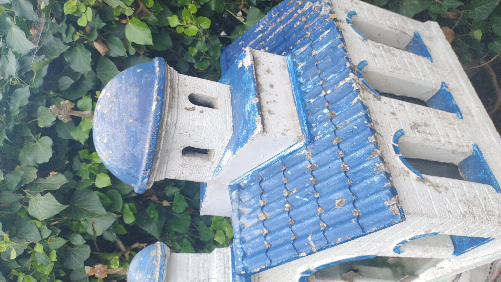 This is a close-up, outdoor, eye-level shot of a white miniature church model with blue-painted details, set against a backdrop of lush green ivy foliage in Νέα Κερασιά, Greece. The model features a blue tiled roof with white trim, and several white walls with arched blue window openings. Above the main structure, two white tiered towers are topped with blue domes. The larger dome on the right has a weathered, antique-looking brown cross affixed to its peak. The smaller dome on the left, partially visible, also has a similar brown cross on top. The paint on the model is somewhat chipped and aged, suggesting it has been exposed to the elements. The ivy leaves in the background are various shades of green, some slightly browned, indicating natural outdoor conditions. The lighting suggests it is daytime, likely overcast or in shade, given the soft light and absence of strong shadows.