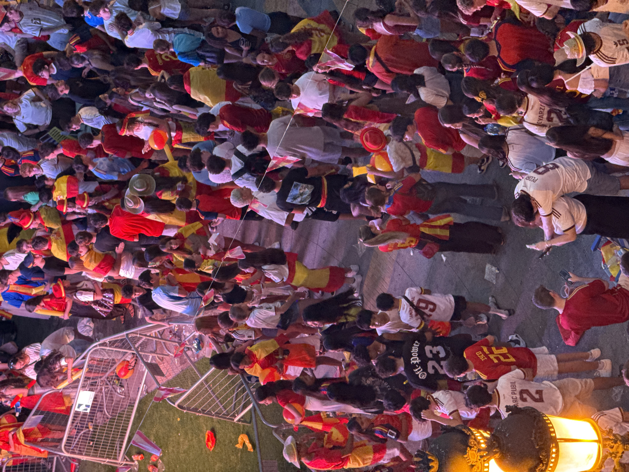 The image shows a crowded outdoor scene, likely a street or plaza, in Las Rozas de Madrid, Spain, as indicated by the prevalence of people wearing Spanish national colors (red and yellow) and Spanish flags. A large group of people, predominantly young adults and teenagers, are gathered, many appearing to be fans or attendees of an event. They are dressed in casual clothing, with a significant number wearing jerseys or t-shirts in red and yellow. Some are holding flags or wearing them as capes. A metal crowd barrier is visible on the left, suggesting a designated area or walkway. The ground is paved, with some litter scattered around. In the foreground, a decorative lamppost with an illuminated globe is on the left. The overall atmosphere appears to be festive and energetic, with people engaged in conversation, holding phones, and interacting with each other. The lighting suggests it is evening or nighttime.