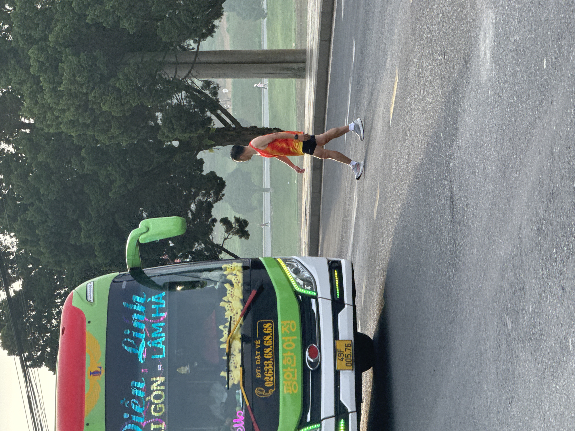 A man in a red and yellow tank top and black shorts is walking across a road in front of a green and white bus. The bus has Vietnamese text and a phone number displayed on its windshield. The man is wearing running shoes and a watch. The background shows trees and a utility pole. The road appears to be a street in the city of Phường Xuân Hương - Đà Lạt, Vietnam.