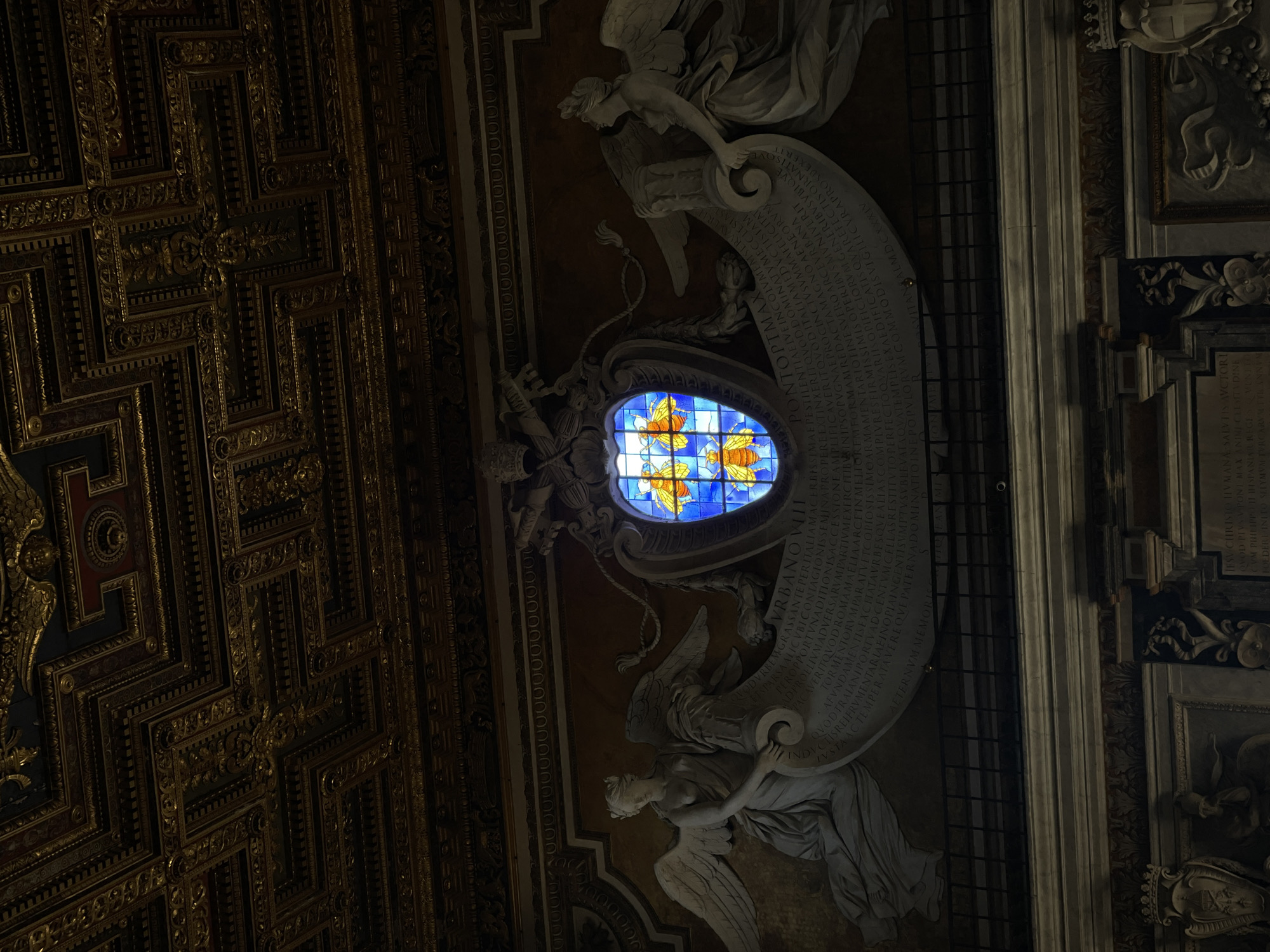 The image captures an ornate interior space within a location in Rome, Italy. The frame is predominantly occupied by a dark, highly detailed, coffered ceiling with gold accents. Moving towards the right side of the image, there is a light-colored wall with sculptures. These include two statues of angels flanking a large, curved inscription or memorial. Positioned at the center between the angel statues is an oval-shaped stained-glass window. This window features a blue background with orange and yellow butterfly or bee motifs. A plaque with text is to the right of the composition.