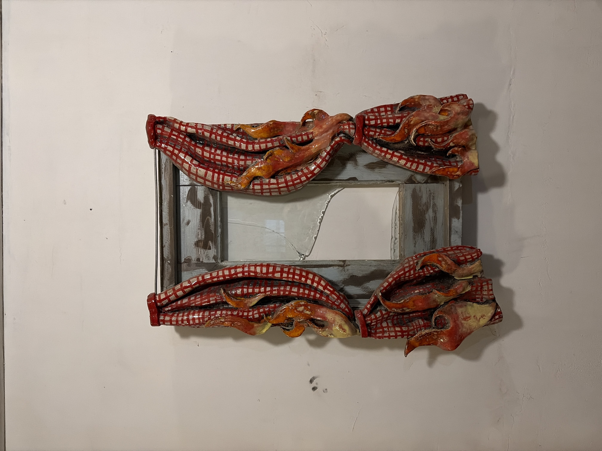 A framed mirror, possibly a piece of art, is mounted on a white wall in Il'inskiy Pogost, Russia. The mirror itself is cracked. The frame is decorated with what appear to be sculpted red and white checkered fabric draped over fiery, orange and yellow elements. There are four such draped and fiery sections adorning the frame, two on each side. The wall has some minor imperfections and shadows.