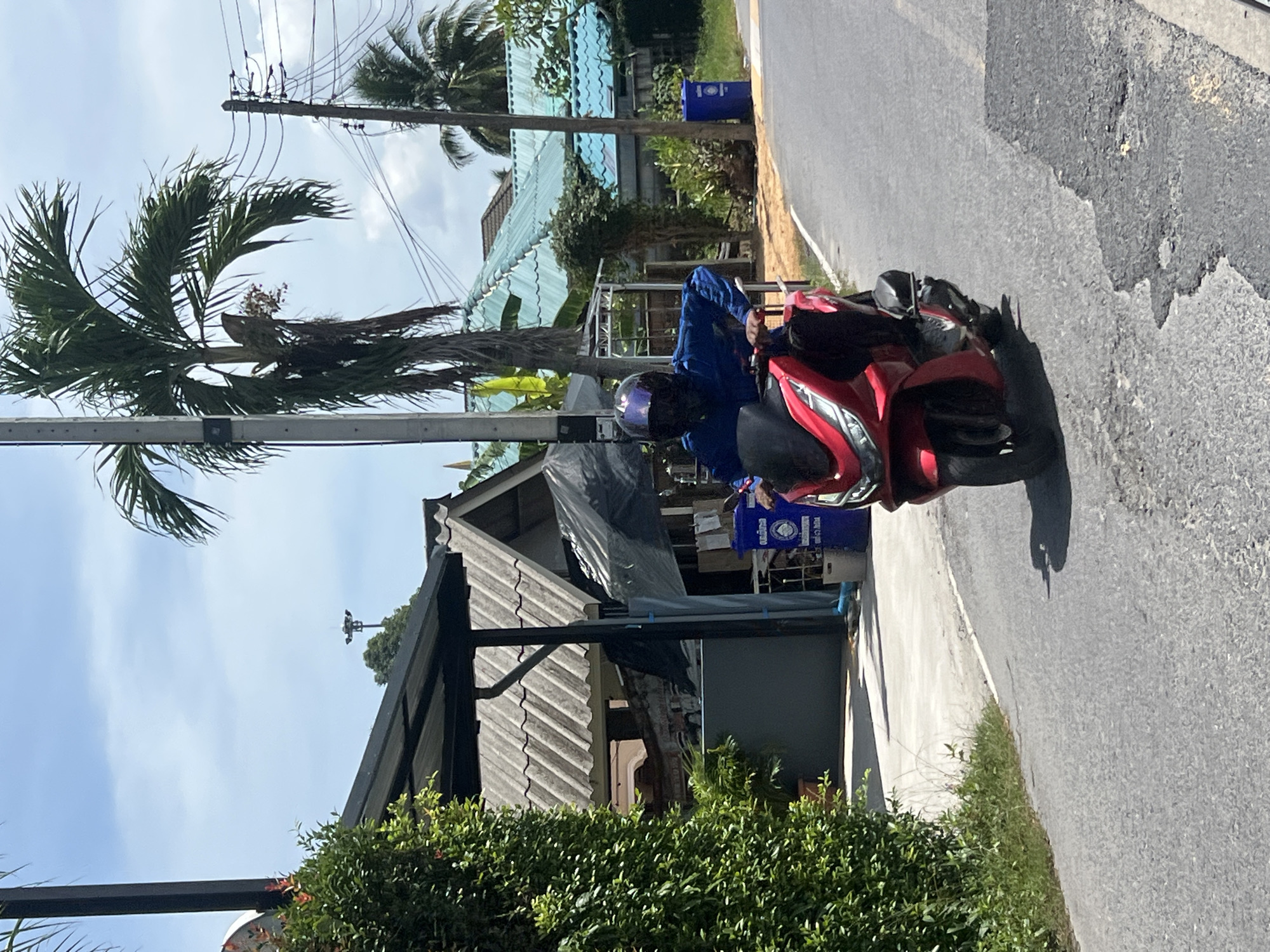A person wearing a blue long-sleeved shirt and a dark helmet is riding a red motorcycle on a paved road. The motorcycle has a black seat and silver handlebars. The person is positioned in the center of the frame, moving towards the viewer. The environment is tropical, with palm trees, lush green foliage, and buildings with gray tiled roofs. In the background, there are utility poles with wires and blue trash bins. The road surface shows signs of wear and tear, with patches of exposed asphalt. The sky is partly cloudy with a bright, hazy sun. The overall impression is of a casual, everyday scene in a warm climate. This scene is located in Choeng Thale, Thailand, a sub-district known for its coastal resorts and vibrant local life.