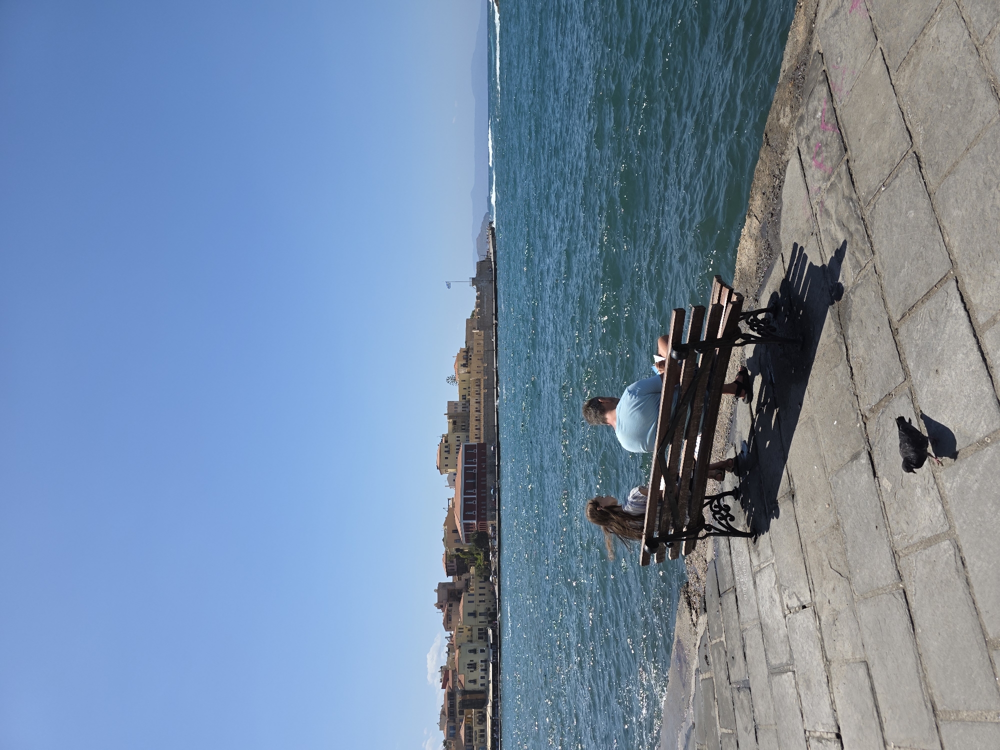A wide shot captures a sunny day in Chania, Greece, with two people sitting on a wooden bench overlooking the Aegean Sea. The sea is a deep blue with whitecaps, and the sky is clear. In the distance, the city's waterfront buildings, painted in shades of yellow and terracotta, are visible. A Greek flag flies from a prominent building. The foreground shows a stone-paved walkway and a single pigeon foraging. The people on the bench, a man and a younger female, appear to be engaged with their phones, with the man wearing a light blue t-shirt and shorts, and the girl a striped top. The bench has ornate metal legs. The overall atmosphere is one of peaceful relaxation by the sea.