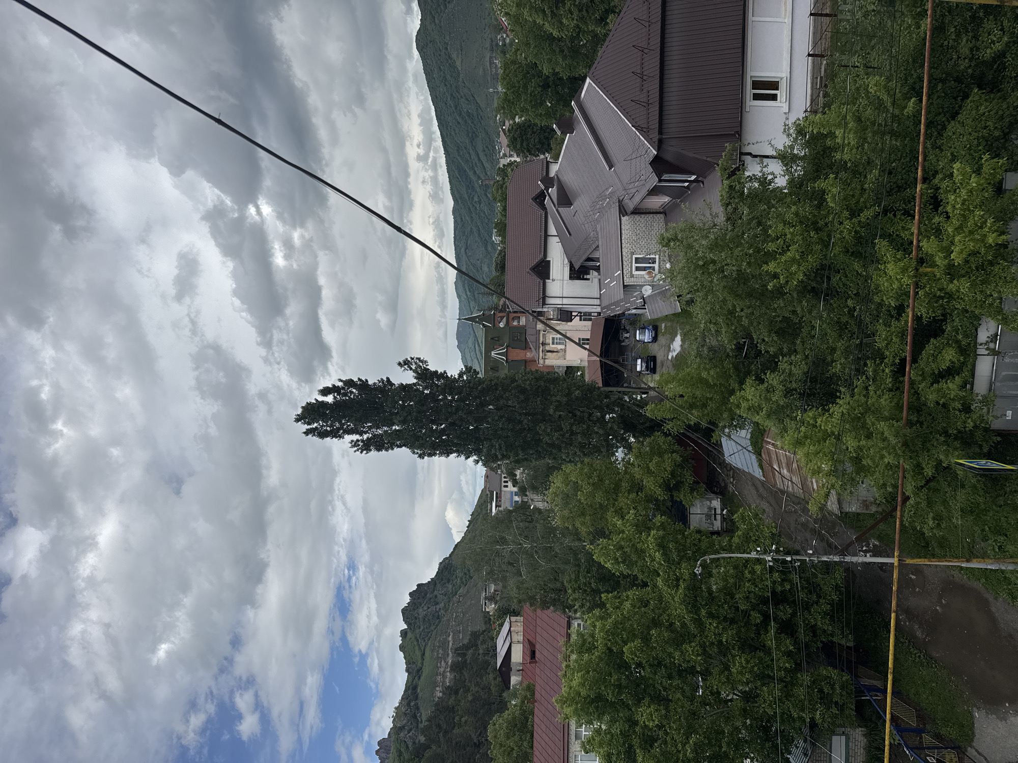 The image depicts an elevated view of a street in Karachayevsk, Russia, on a cloudy day. The foreground includes power lines and the upper parts of trees with green foliage. Several buildings with varying roof colors (brown, gray, red) are visible, along with a few cars parked on the street. In the background, there is a mountain range, partly obscured by clouds. The sky is overcast with a mix of gray and white clouds, with a hint of blue visible in the lower left corner. A tall, narrow tree stands prominently in the middle ground. A street lamp is visible in the bottom right corner of the frame.