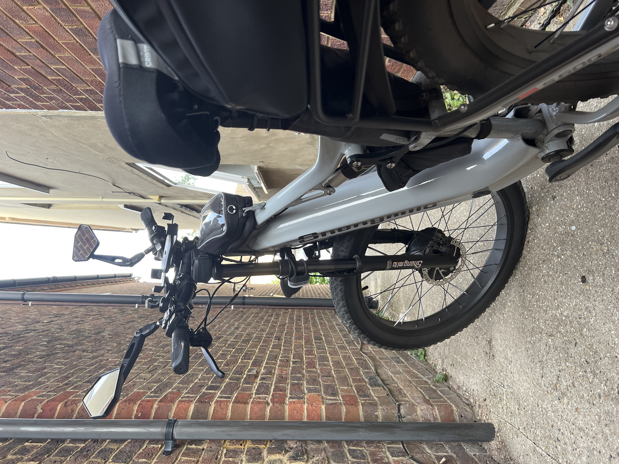 This media file shows a close-up, angled view of an electric bicycle, predominantly the rear wheel, frame, and handlebars, mounted on a brick wall in an outdoor setting. The bicycle is light grey with black accents, including the tires, saddlebag, and handlebar grips. The rear wheel is large and appears to be a fat tire, with a black rim and spokes. The electric motor is visible within the hub of the rear wheel. Text on the motor housing reads "Shengmilo". A black saddlebag is attached to the rear rack above the wheel. The handlebars are equipped with two black mirrors, one on each side, and various controls.
The scene is set outdoors, with a brick wall forming the backdrop. The ground is paved with concrete. The lighting suggests it is daytime, possibly overcast given the diffused light. The overall impression is of a parked bicycle, possibly leaning against the wall or being stored. There are no people visible in the image. The setting appears to be a residential area or possibly a bike storage area. The location context of Guildford, United Kingdom, implies this is a typical urban or suburban environment. The image is captured from a low angle, looking up towards the bicycle.