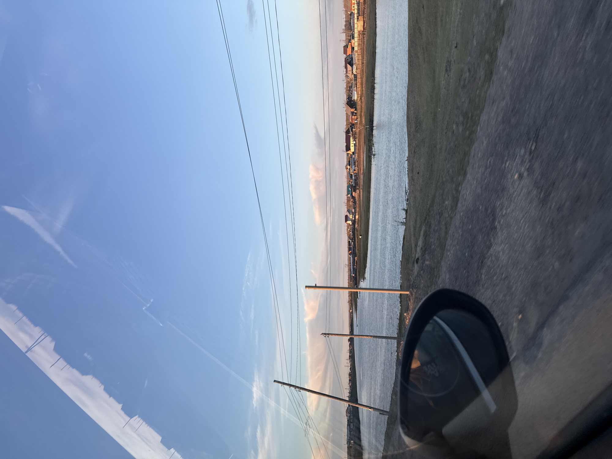 The image appears to be taken from inside a vehicle, possibly a car, with a view of Syrdakh, Russia. The side mirror is visible in the foreground. Outside the vehicle, there is a body of water (possibly a river or lake) running parallel to a line of buildings, likely residences, along the shoreline. Overhead, there are power lines suspended by utility poles. The sky is a gradient of blue, suggesting either sunrise or sunset. The ground visible to the right of the water appears to be vegetation or a grassy area, bordered by what looks like a road surface. The reflection in the car's window may show similar elements to the outside scene. The photograph is oriented sideways.