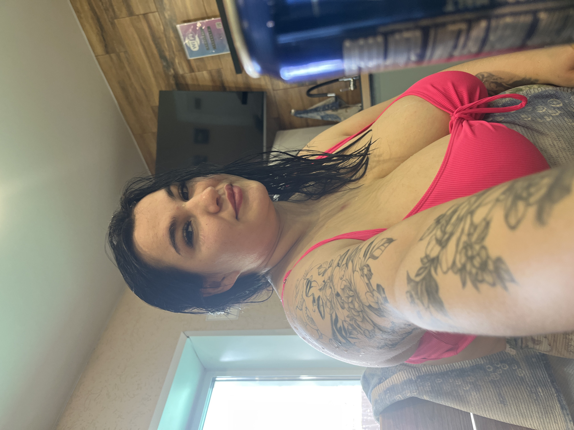 The image shows a woman with dark, wet hair wearing a pink bikini. She has tattoos on her left arm. The background includes a light-colored wall, a window, a television, and a card with the word "WiFi" printed on it. The location may be in Chaikovsky, Russia.