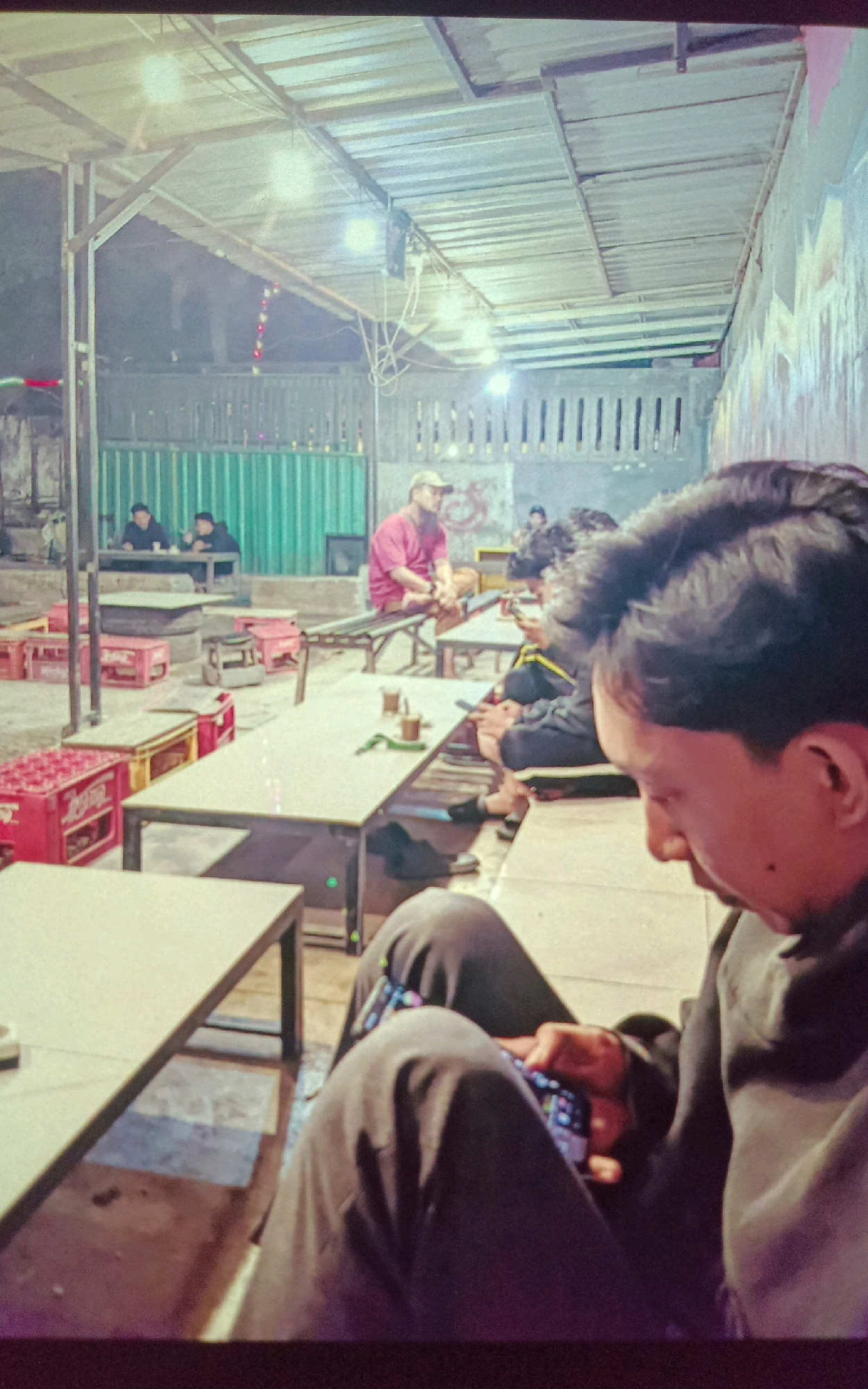 The image depicts an outdoor seating area of a cafe or eatery, likely located in Gampengrejo, Indonesia, given the context provided. The setting appears to be a casual, open-air establishment with a corrugated metal roof supported by metal beams, providing shade. The ground is paved with concrete.
In the foreground, a person is seated, facing away from the camera and looking down at a mobile phone. This individual is wearing dark pants and a dark hooded jacket. Their legs are crossed, and the phone is held in their hands. The background reveals other patrons and elements of the establishment.
Several tables and benches are arranged throughout the space. One table in the mid-ground has at least two cups on it, suggesting drinks were served. To the left of this table, red crates are stacked, some of which appear to be beverage crates. Further in the background, more tables are visible, with people seated at them. One person in the mid-background is wearing a bright red shirt and a cap, appearing to be engaged in conversation or observing something. Another group of people are seated at a table on the far left.
The overall atmosphere suggests a relaxed, communal gathering space, possibly an informal eatery or a coffee shop. The lighting appears to be artificial, likely from overhead lamps, contributing to a somewhat dim, evening ambiance. The walls behind some of the seating areas feature some graffiti or painted designs. The presence of green corrugated metal panels suggests a partition or part of the building's structure.