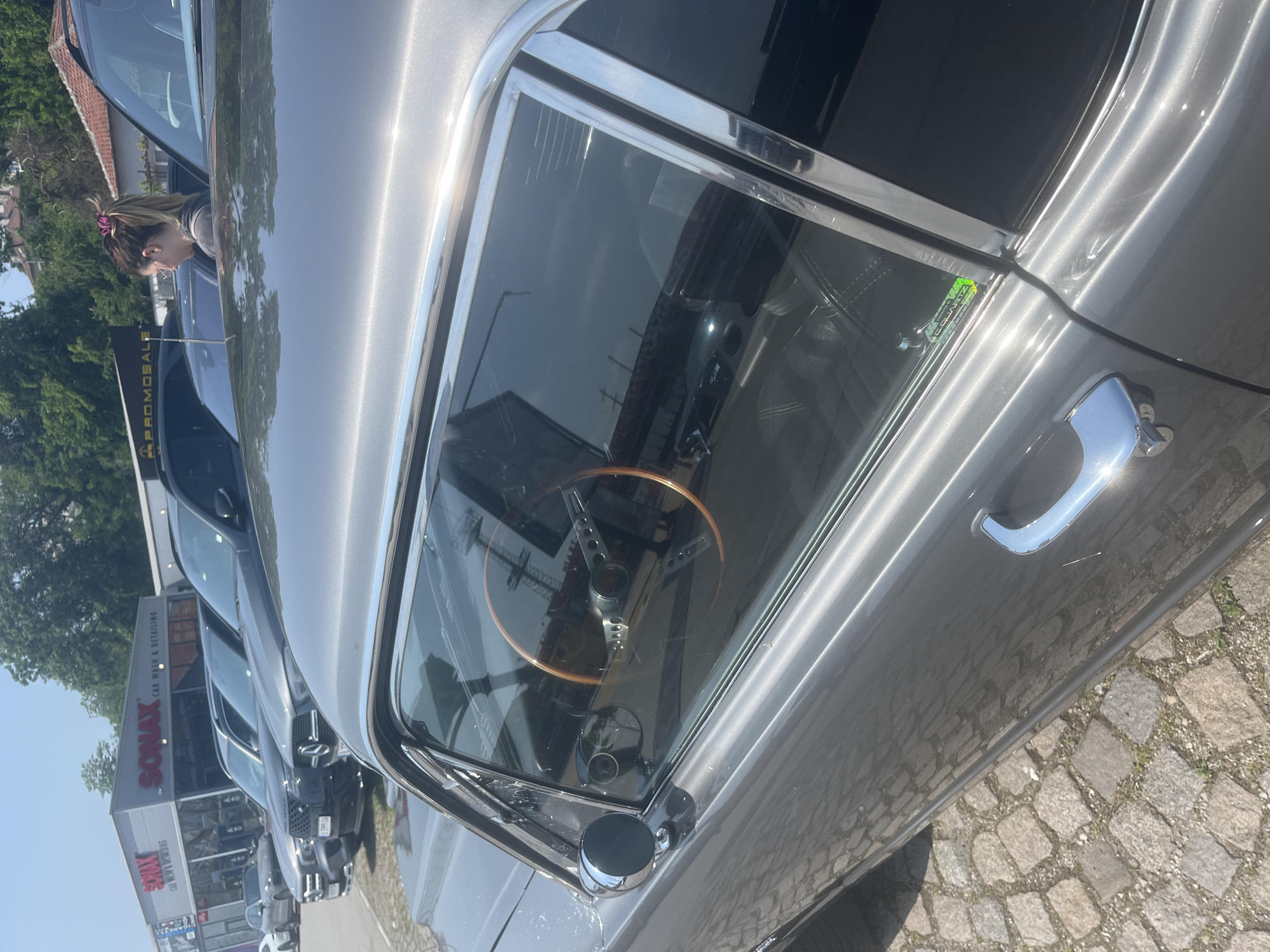 The image captures a sunny daytime scene at a car wash or detailing facility in Varna, Bulgaria. The primary subject is the side view of a gleaming grey vintage or classic car, parked on a cobblestone surface. Its chrome window trim, side mirror, and door handle reflect the strong sunlight. Through the car's window, a distinctive wooden steering wheel with metal spokes is clearly visible.
In the midground, a person, likely a woman with light brown hair tied in a ponytail with a pink scrunchie, leans into the open window of a dark-colored modern car, looking towards its interior. Behind this, several other modern vehicles are parked.
The background features a building with prominent red "SONAX CAR WASH & DETAILING" signage, indicating the location's purpose. A black flag with white "PROMOSALE" text is also visible. The sky is clear and blue. A small, reflective green sticker on the classic car's side window appears to read "CERAMIC QUARTZ COATING," suggesting the vehicle may have recently received a detailing treatment. The overall setting implies a car-related event, dealership, or service center.