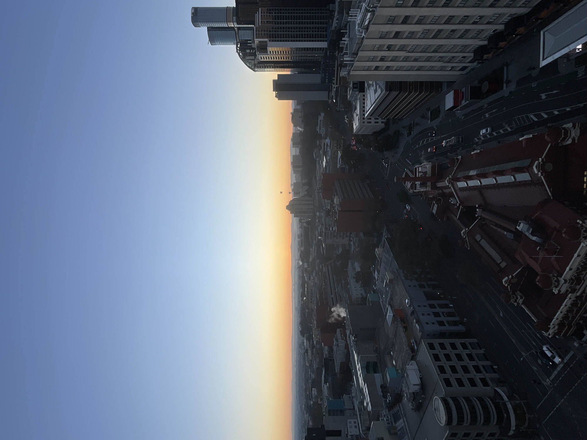 This aerial photograph captures a sunrise over the city of Melbourne, Australia. The sky transitions from a deep blue at the top to a soft orange and yellow gradient near the horizon, indicating the early morning. A thin layer of haze is visible over the city, obscuring distant features.
In the foreground, several buildings of varying architectural styles are visible. On the right, a cluster of modern, tall glass skyscrapers dominates the skyline, reflecting the dawn light. To the left of these, lower-rise buildings, some with older facades, fill the middle ground. A prominent building with a red, ornate roof is situated in the lower central part of the frame, suggesting a historic structure. Several roads are visible winding through the city, with minimal traffic in the early morning light. Some cars are present on the roads and parked in designated areas.
In the distance, several hot air balloons are visible, adding a point of interest to the scene. Smoke or steam is rising from some of the buildings, possibly from industrial activity or heating systems. The overall impression is one of a city waking up to a new day, bathed in the warm glow of the rising sun. The perspective is from a high vantage point, offering a sweeping view of the urban landscape.