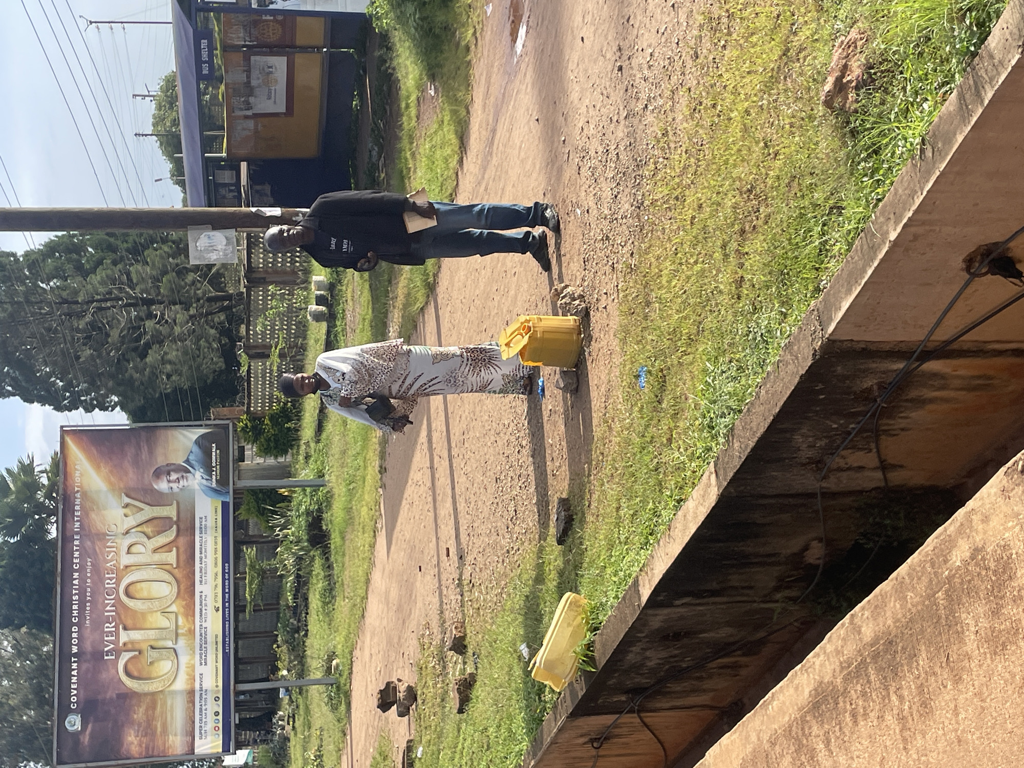 In Jos, Nigeria, two individuals stand on a dirt road near a bus shelter. A large billboard with the text "COVENANT WORD CHRISTIAN CENTRE INTERNATIONAL invites you to enjoy EVER-INCREASING GLORY" is visible in the background. A woman, dressed in a patterned garment, stands to the left, holding a dark bag. To her right, a man in a black jacket and jeans holds a book. A yellow container and several bricks are on the ground. The environment includes grass, trees, and a concrete drainage channel in the foreground.
