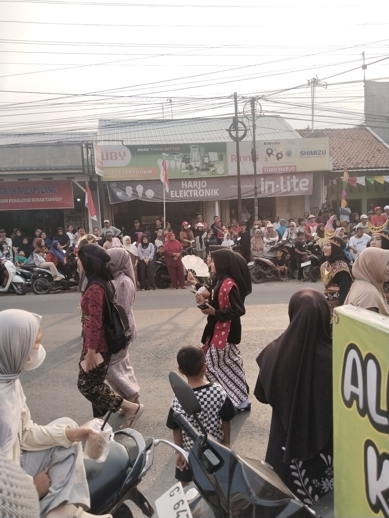 The image captures a street scene in Balapulang Kulon, Indonesia, during what appears to be a cultural event or parade. A diverse crowd of people, many wearing traditional attire, line the street, observing the proceedings. Several motorcycles are parked along the roadside, indicating transportation common in the area. In the background, shops and banners with Indonesian text are visible, suggesting a commercial district. The sky is overcast, and power lines crisscross the frame, characteristic of urban environments in Indonesia. The foreground is occupied by individuals in vibrant clothing, some holding fans, adding to the festive atmosphere. The overall impression is one of community gathering and celebration within a lively Indonesian town.