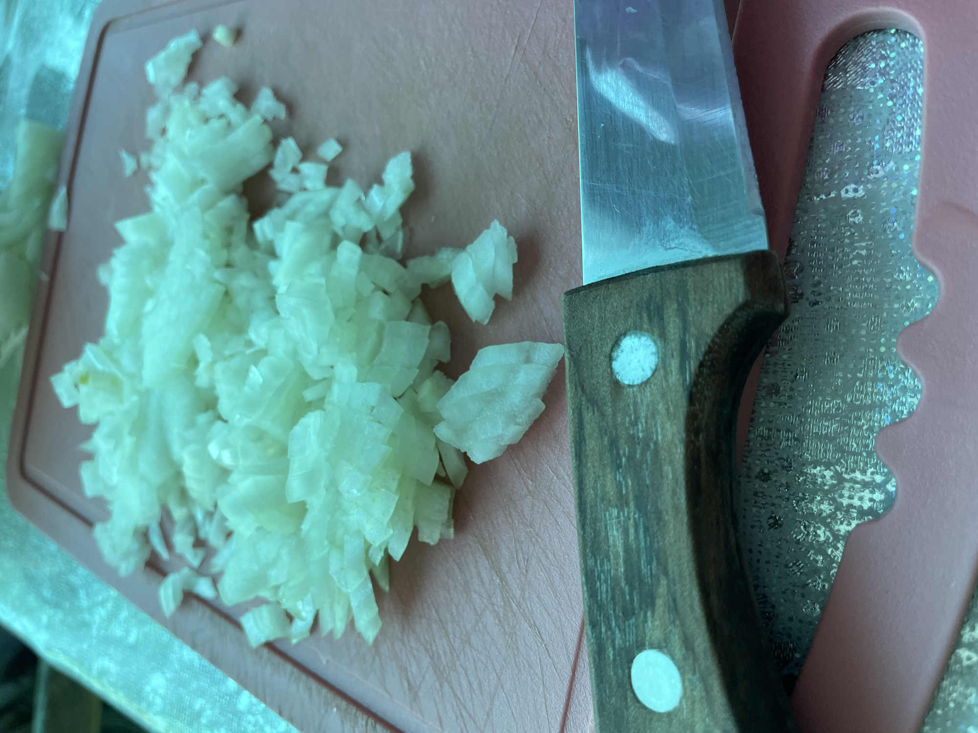 A close-up shot depicts diced onions arranged on a pink cutting board, next to a knife with a wooden handle. A section of another tool with a glittery, silver surface is partially visible near the knife.