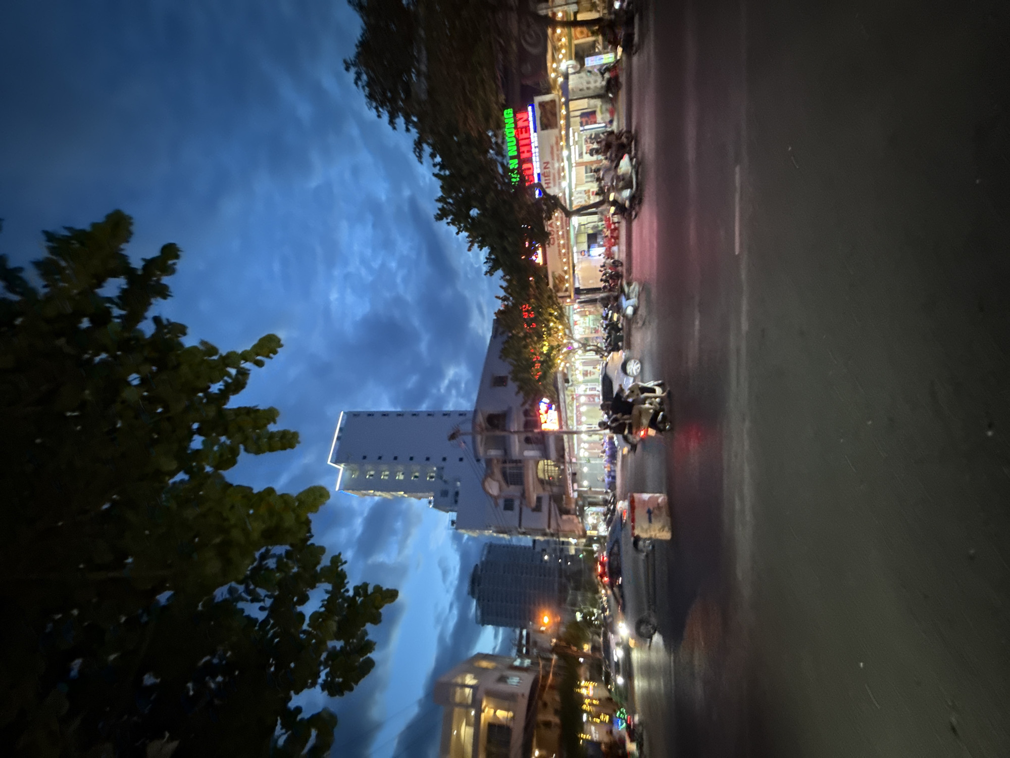 The image depicts a street scene in Nha Trang, Vietnam, at dusk. The sky is a deep blue with dramatic clouds, suggesting twilight. In the foreground, a road is visible with several motorcycles and a few cars. On the right side of the road, there are buildings with illuminated signs, indicating businesses. One sign appears to be in Vietnamese and displays colorful neon lights. Trees with dark foliage are scattered throughout the scene, some obscuring parts of the buildings and sky. The overall atmosphere is that of a bustling urban environment transitioning into the evening.