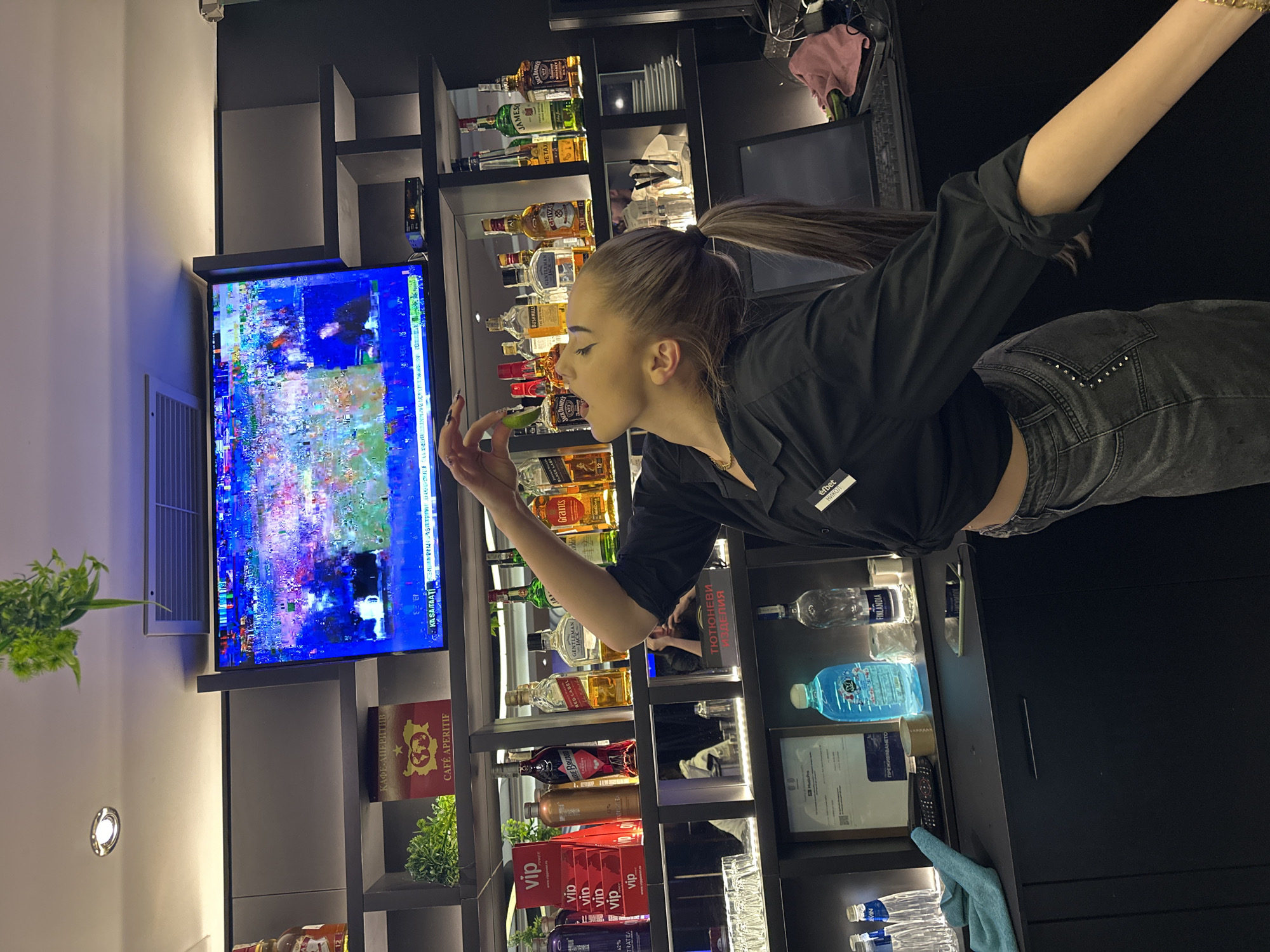 This image shows a young woman with her hair pulled back in a ponytail, wearing a black shirt and grey jeans. She is in a bar or lounge area, standing in front of a shelf stocked with various bottles of alcohol. A television screen in the background displays a distorted image, likely due to a signal issue. The woman is holding a slice of lime up to her mouth, as if to taste it, with a playful expression on her face. The lighting in the scene is dim, creating a cozy atmosphere. Visible text includes brand names on the alcohol bottles like "Jameson," "Grant's," "Red Label," and "Black Label," as well as signage for "Café Aperitif" and "Vip." There are also some bottles of water and what appear to be cigarette products displayed. The overall impression is a snapshot of a bar employee in a moment of playful engagement.