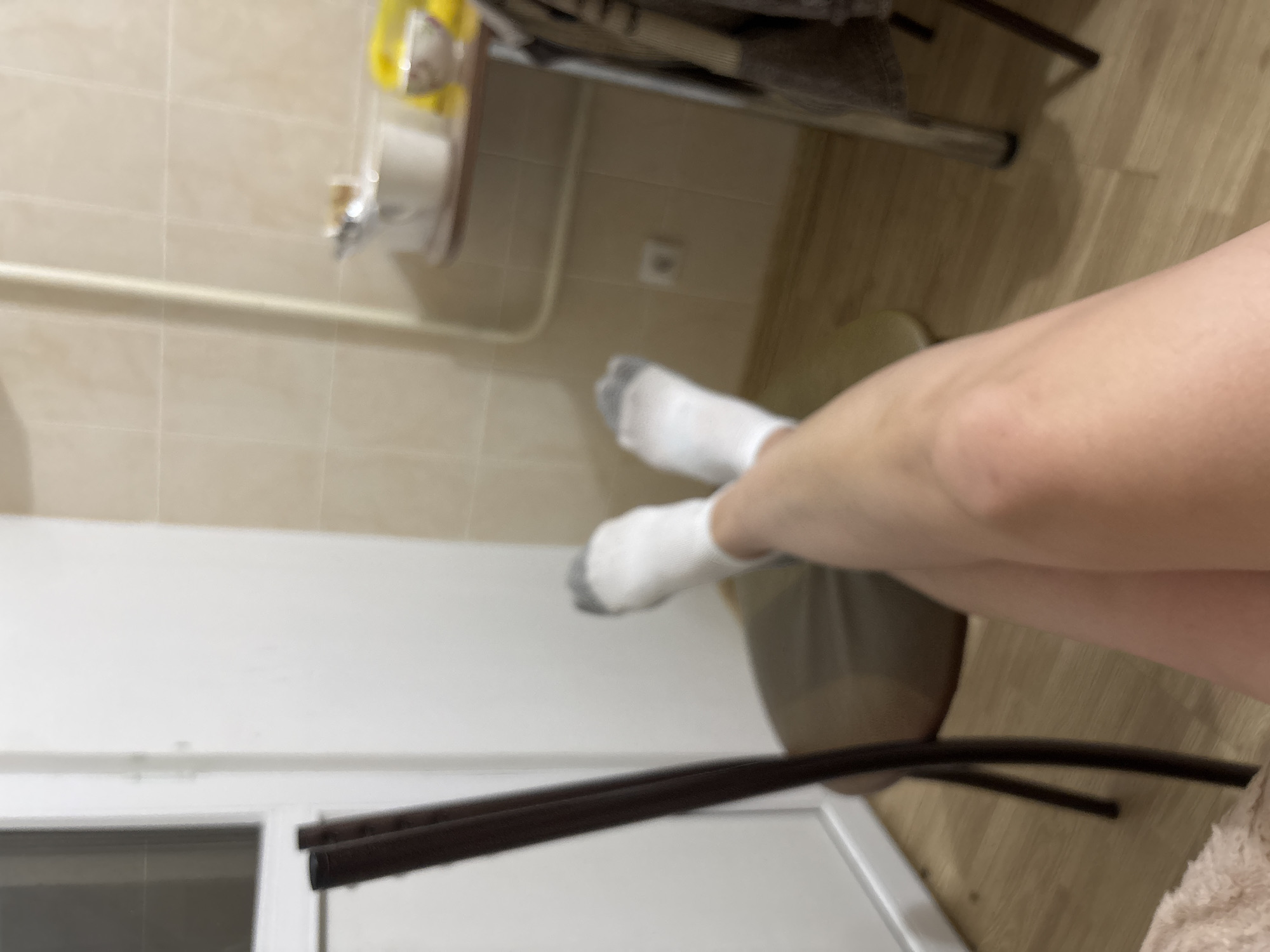 A close-up, slightly angled, indoor shot features a pair of legs wearing white socks. The legs are positioned in the lower right portion of the frame. The background consists of a wall with beige tiles, a towel rack, a metal frame structure, and a white window frame with two brown poles. Based on the interior elements, the location appears to be a bathroom. There is no evidence to support that this image was taken in Sochi, Russia.
