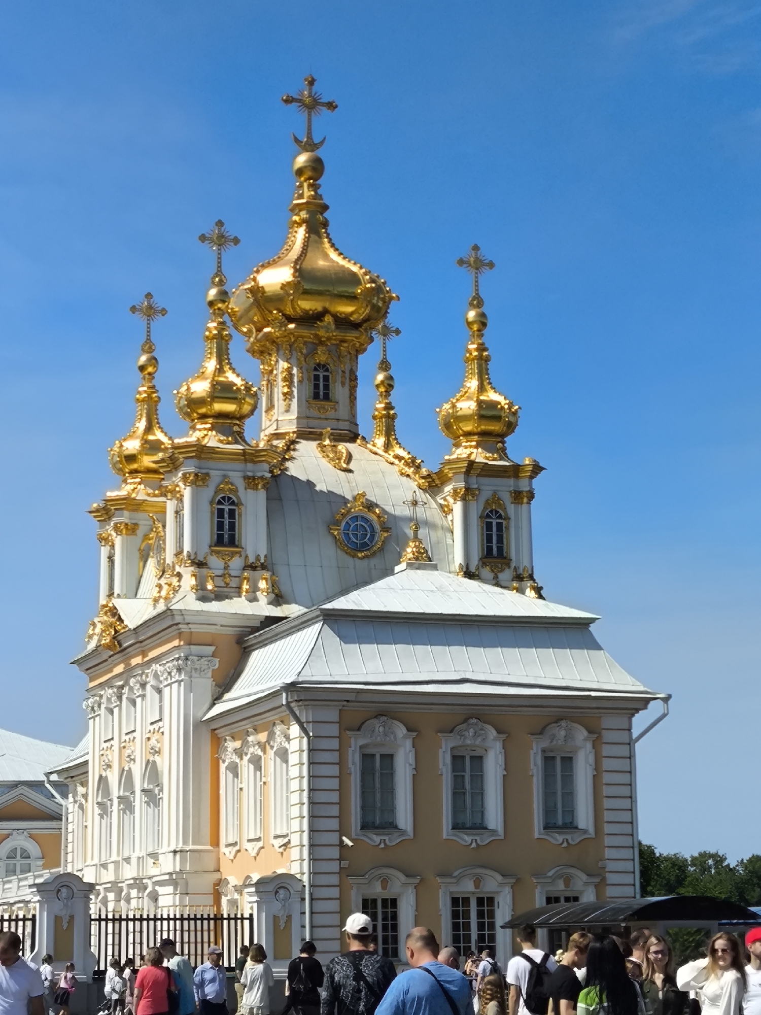 This is a vertical, eye-level shot of the Church of the Ascension in Peterhof, Russia, on a clear, sunny day. The church is a prominent golden-domed structure with white and yellow accents. Several smaller golden domes and ornate crosses adorn its roof. The building features arched windows with white trim and decorative carvings.
In the foreground, a group of people are gathered. Some are walking, while others appear to be observing the church. Among them are individuals wearing casual clothing, including t-shirts, shorts, and hats. A black metal fence runs along the bottom of the frame, separating the people from the church. Lush green trees are visible in the background, contrasting with the bright blue sky.