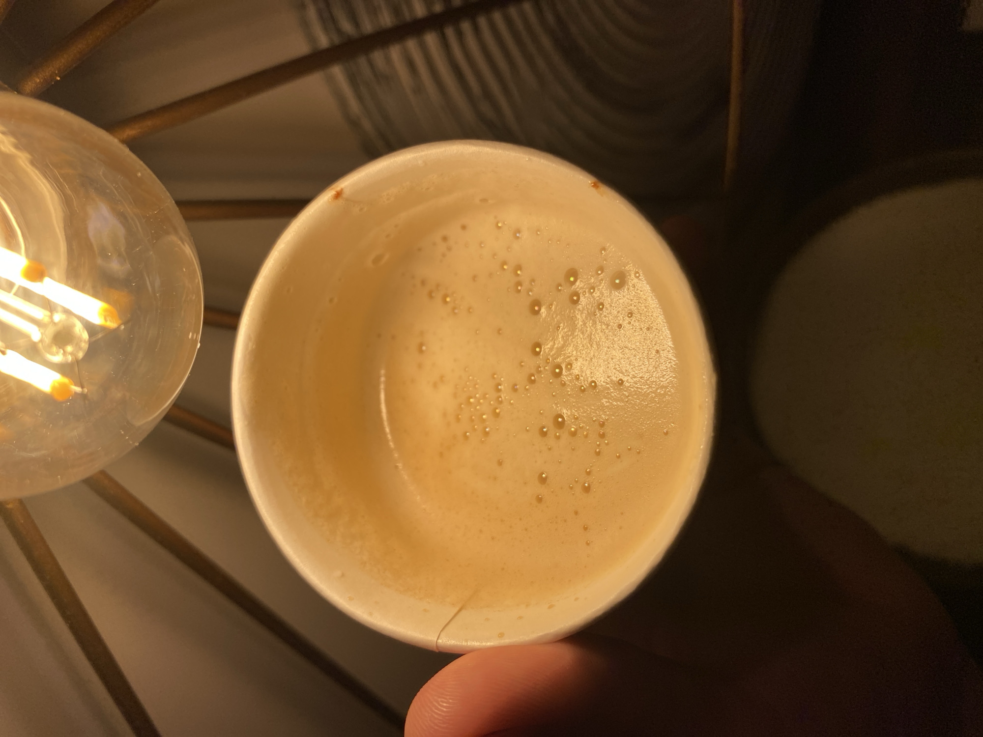 A close-up, top-down view shows a white paper cup filled with foamy, light brown liquid, likely coffee or a similar beverage. The liquid has a frothy surface with small bubbles. A person's thumb and index finger are visible on the left side of the frame, holding the cup. Above the cup, a clear glass light bulb with glowing filament is illuminated, casting a warm light on the scene. The background is dark and appears to be part of a lamp fixture with metal struts and a textured surface. The setting is indoors, with the lighting suggesting an evening or dimly lit environment. There is no information in the image to determine the location in Votkinsk, Russia.