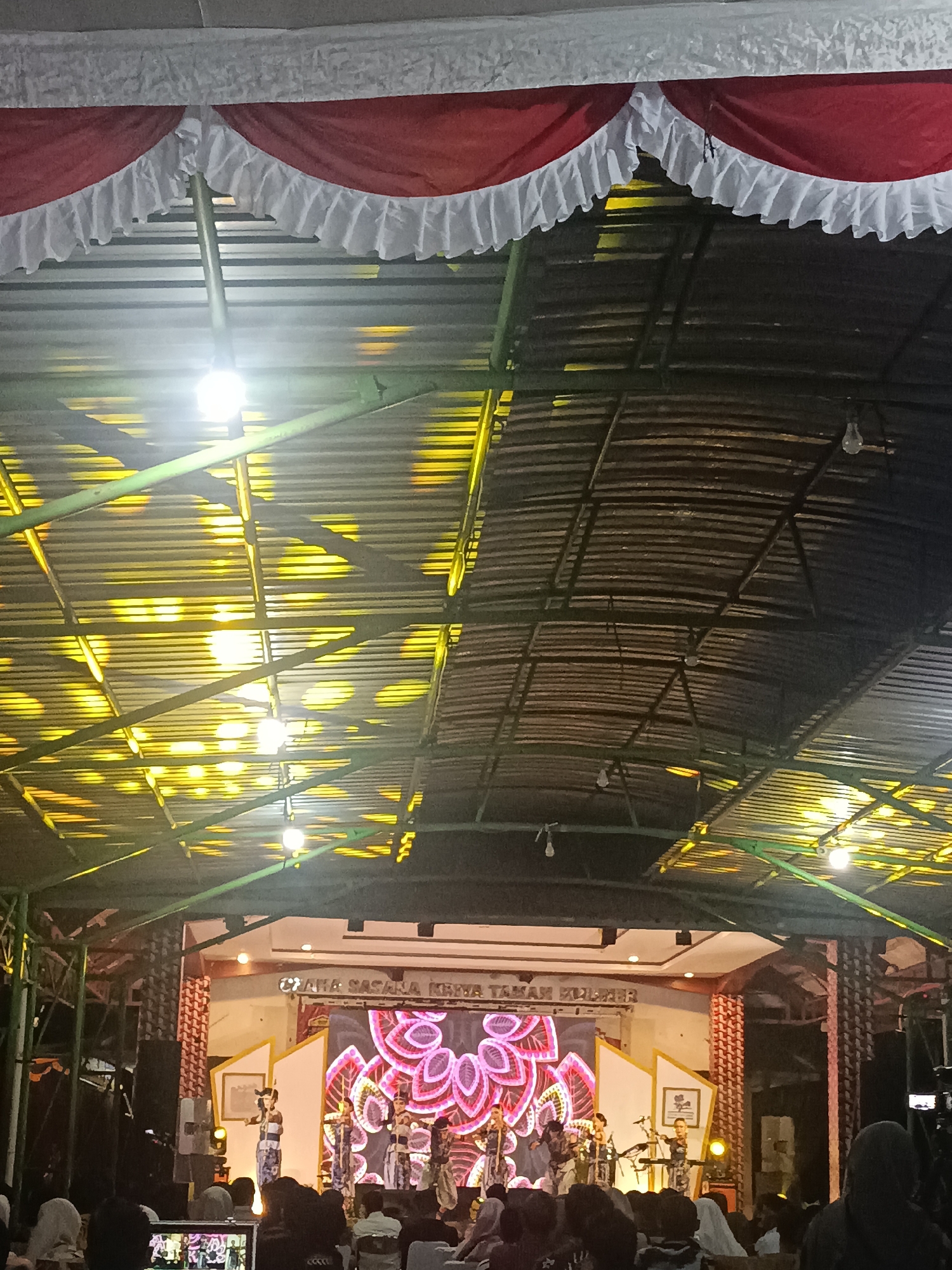 The image depicts an indoor performance stage, likely in Condongcatur, Indonesia, with a decorated roof featuring red and white ruffled fabric. The stage itself is illuminated by bright yellow lights casting patterned shadows on the corrugated metal ceiling. A large screen at the back of the stage displays a vibrant pink floral design. Several performers, dressed in traditional attire, are on stage, some holding musical instruments. An audience, mostly out of focus and partially visible in the foreground, is seated, suggesting a cultural event or performance. The overall atmosphere appears to be festive and cultural.