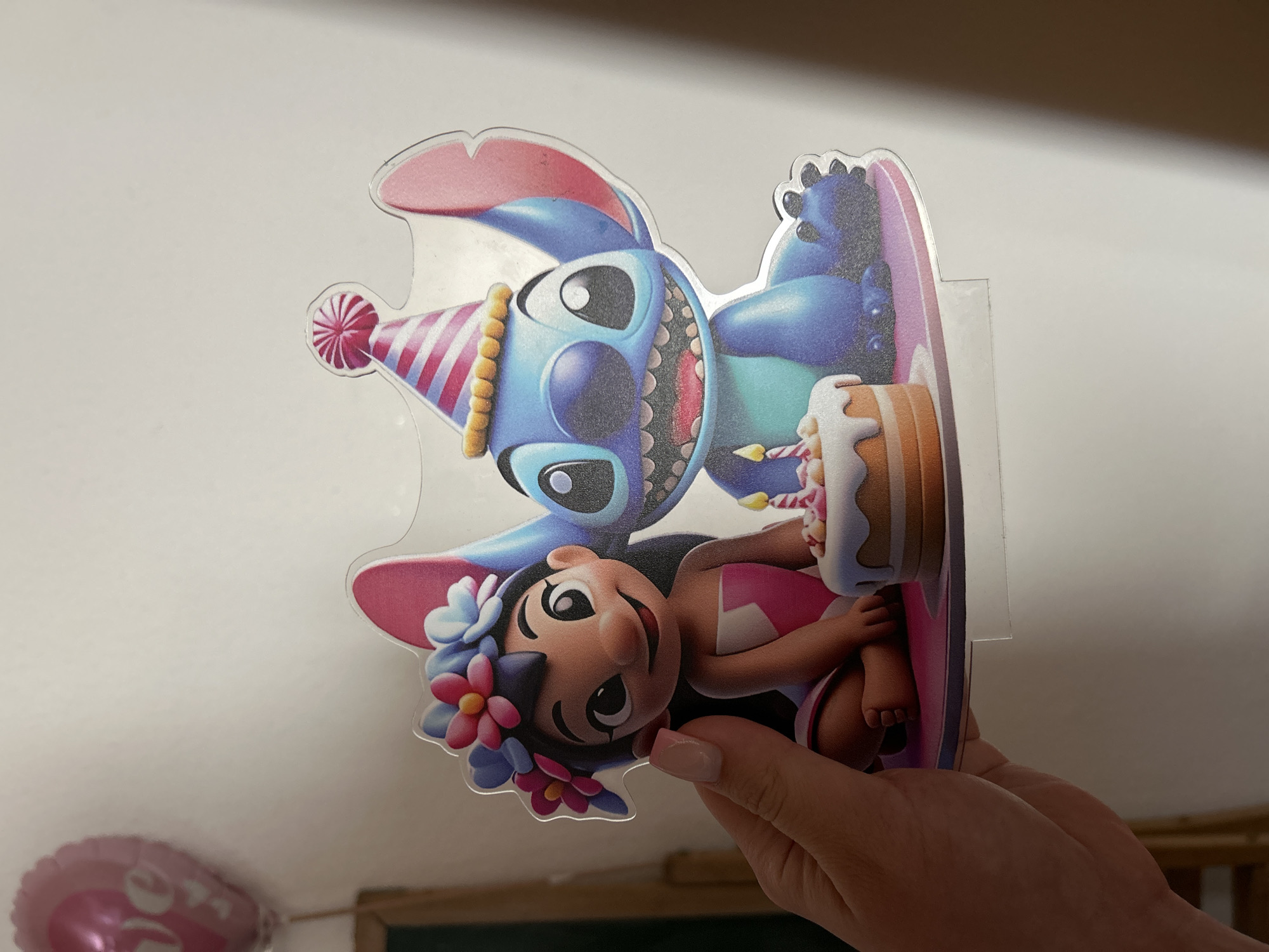 This image, taken in the city of Waldbröl, Germany, depicts a left hand holding a laminated cardboard cutout featuring the Disney characters Lilo and Stitch. The cutout portrays Lilo, a young girl with dark hair and a flower crown of blue and pink blossoms, smiling broadly and wearing a pink top and skirt. To her right sits Stitch, a blue alien, also smiling broadly and wearing a pink and white striped party hat. A small, two-tiered birthday cake with white frosting, pink and white decorations, and two lit candles is positioned in front of the characters. Both Lilo and Stitch are depicted seated on a pink and purple circular base, suggesting a celebratory theme. The hand holding the cutout has manicured fingernails with a light polish. The background consists of a plain, light-colored wall. In the upper-left background, a blurry pink heart-shaped balloon with indistinct white text is partially visible, along with a faint wooden edge of an unknown object below it. The lighting suggests an indoor setting.