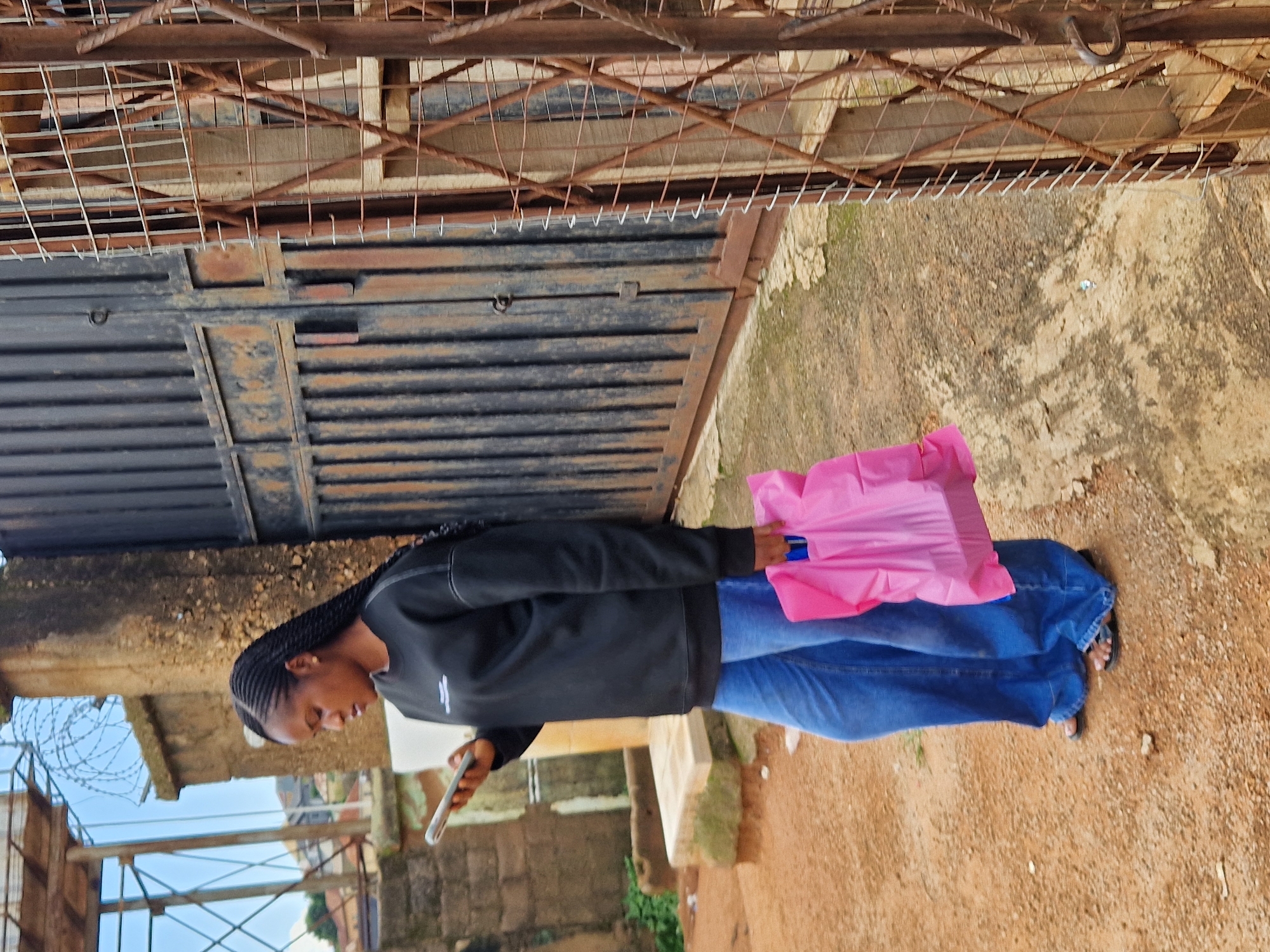 In the city of Unknown, Unknown, a young woman with braided hair stands on a dirt path, looking at her phone. She is wearing a black sweatshirt and blue wide-leg jeans, with black flip-flops. In her right hand, she carries a bright pink plastic shopping bag. Behind her, a large, dark metal gate with vertical slats stands partially open. To the right of the gate, a rusted metal fence with diamond-shaped mesh is visible. The ground is composed of loose dirt and small stones. In the background, a brick wall and a structure with barbed wire can be seen. The sky is overcast.