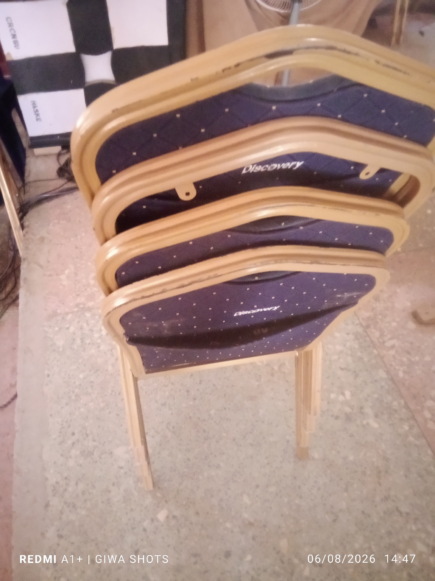 The image shows a stack of four chairs with light wood frames and dark blue seats with a white polka dot pattern. The word "Discovery" is visible in white lettering on the seats of the lower two chairs. The chairs are stacked on a light-colored, speckled floor. In the background, a black and white checkered board is partially visible, along with a white sign that has black lettering, some of which reads "HASKE" and "CRCNBU". The scene appears to be indoors, possibly in a storage area or event venue. The image also has a timestamp and text overlay at the bottom, reading "REDMI A1+ | GIWA SHOTS" and "06/08/2026 14:47".