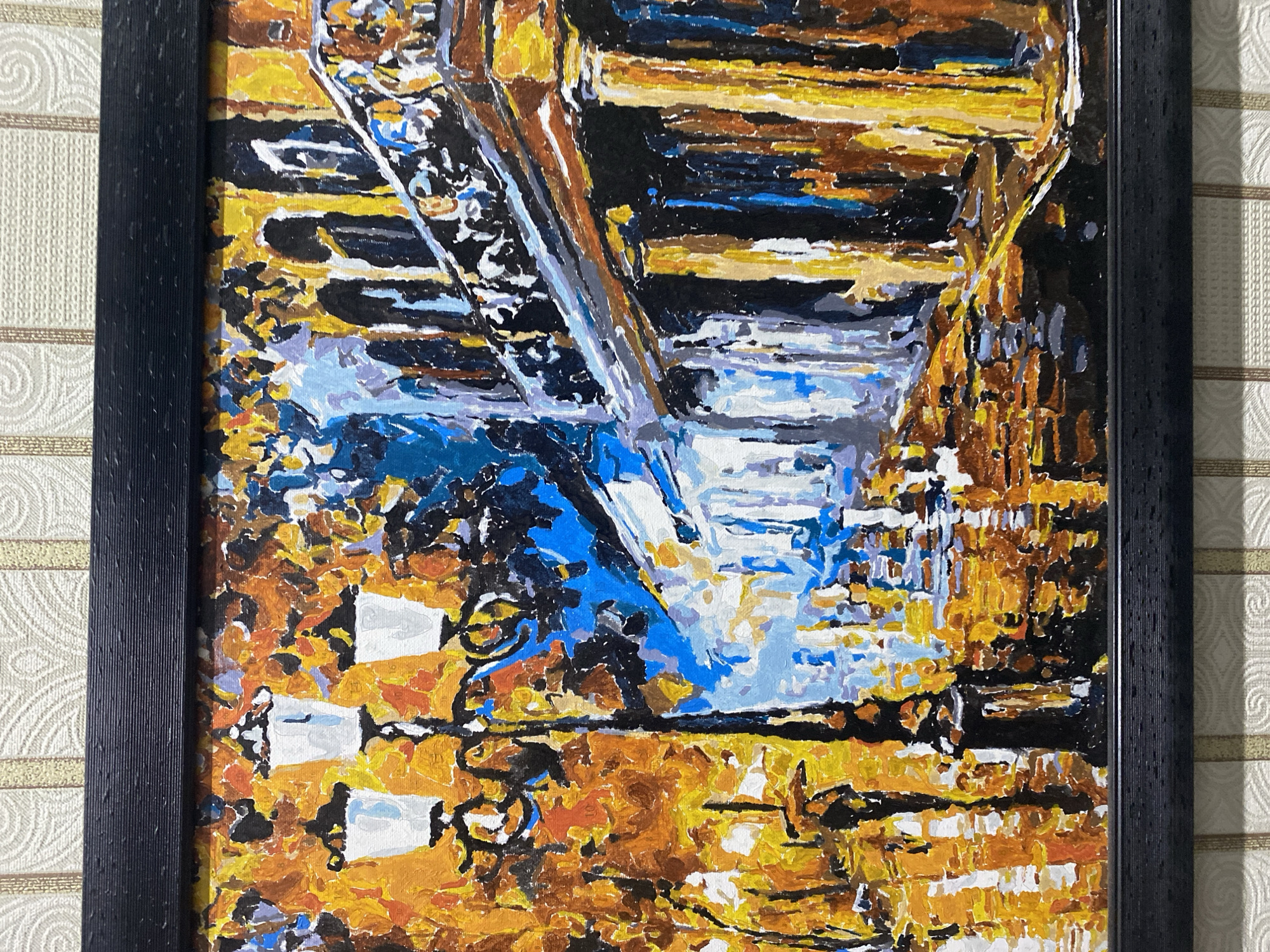 The image depicts a thick impasto oil painting of a street scene, likely in autumn given the dominant warm tones of orange and yellow. The style is impressionistic, with visible brushstrokes and a focus on light and color rather than sharp detail.
In the foreground, a lamppost with three white lanterns stands prominently on the left. The street appears wet, reflecting the warm colors of the surrounding trees and the bright lights. A lone figure holding an umbrella is walking away from the viewer on the left side of the street, partially obscured by the lamppost and foliage.
To the right, the facade of a building is depicted. It features arches and columns, rendered in dark blues, blacks, and yellows, with a decorative railing on an upper level. The reflection of the building and its lights is also visible on the wet street. The overall impression is of a city street illuminated by streetlights on a damp, possibly rainy, evening or morning during the fall season.
There is no discernible information or visual cues in the painting that would identify the location as Chernogorsk, Russia, or any other specific city. The description is based solely on the visual content of the artwork.