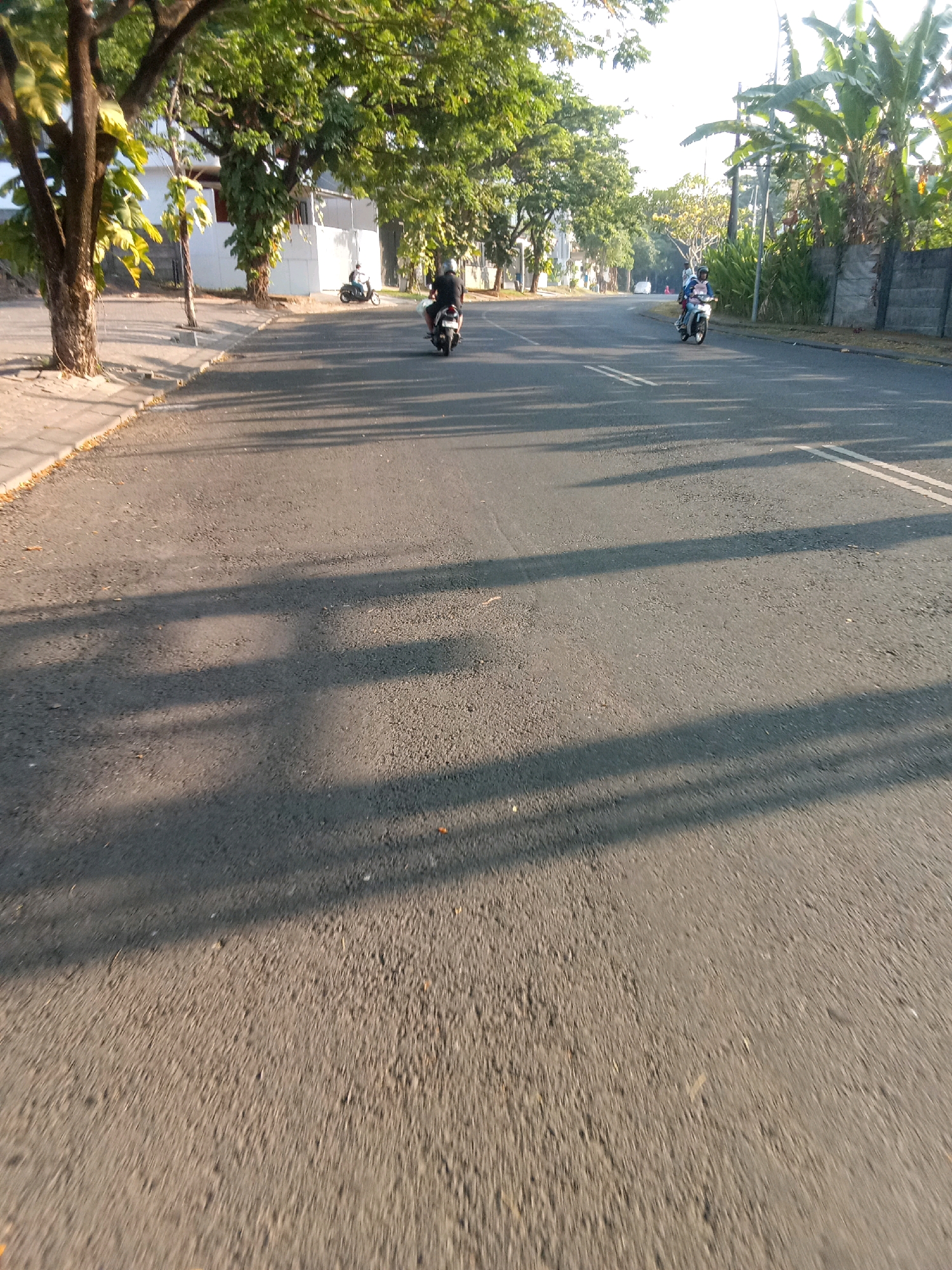 This is a photograph of a street scene in Jember, Indonesia. The road is paved and has white lane markings. Trees line the left side of the road, and a wall borders the right side. Several people are riding motorcycles on the road. The sun is shining, casting long shadows across the asphalt. The overall impression is one of a quiet, sunny day in a residential or suburban area.