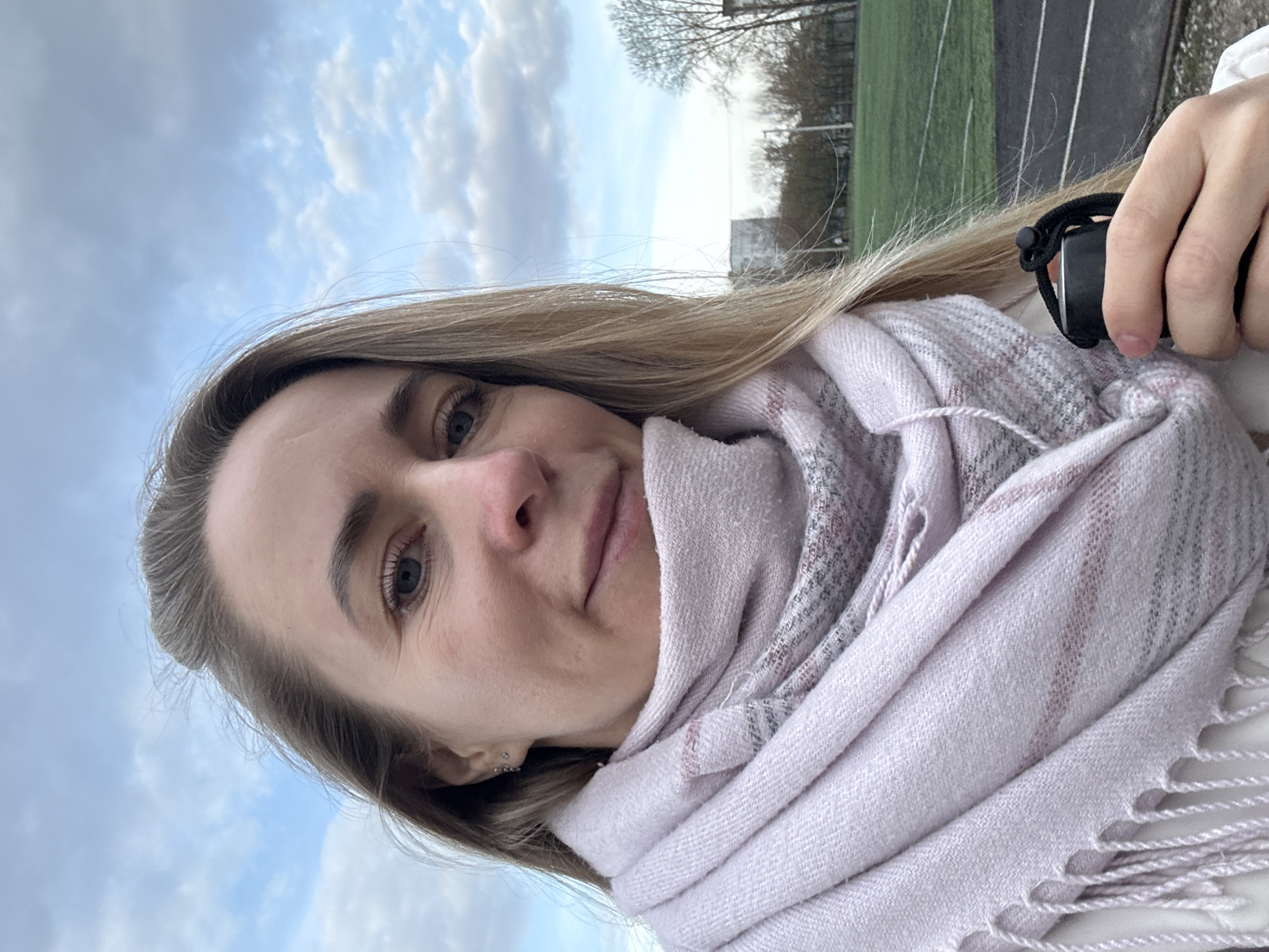 A selfie taken in Chaikovsky, Russia, features a fair-skinned woman with blonde hair and blue eyes, wearing a light purple and grey scarf. The background shows a cloudy sky, trees, and a running track, which indicates a park or sports facility. The woman is holding a black object in her hand.