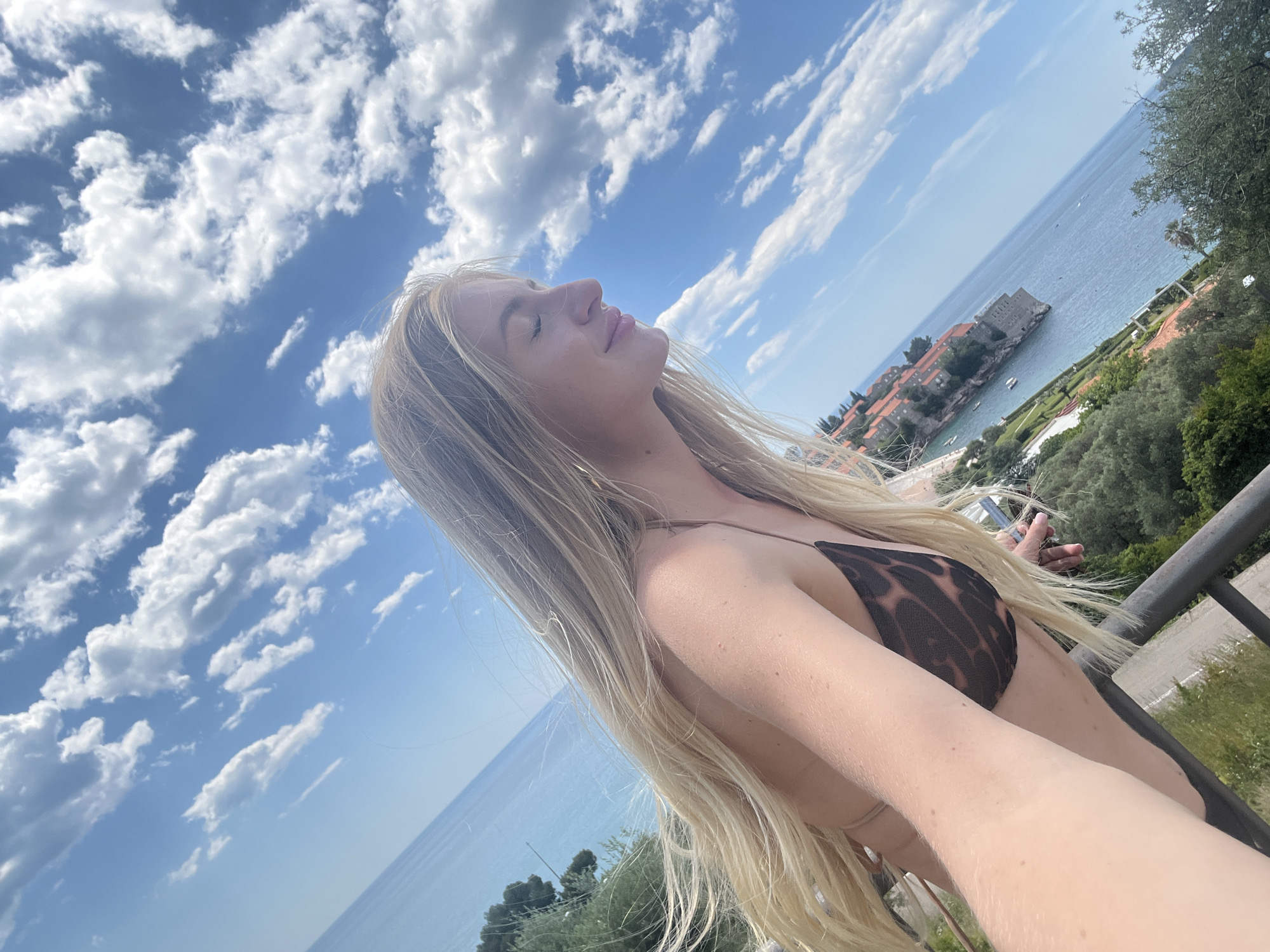 A woman with long blonde hair stands on a balcony with a metal railing on a sunny day in Sveti Stefan, Montenegro. She is wearing a brown leopard print bikini and has her eyes closed, her face turned towards the sky. Behind her is a view of the Adriatic Sea, a cloudy blue sky, and the islet of Sveti Stefan. There are green trees and vegetation visible around the railings and the town of Sveti Stefan is visible in the background.