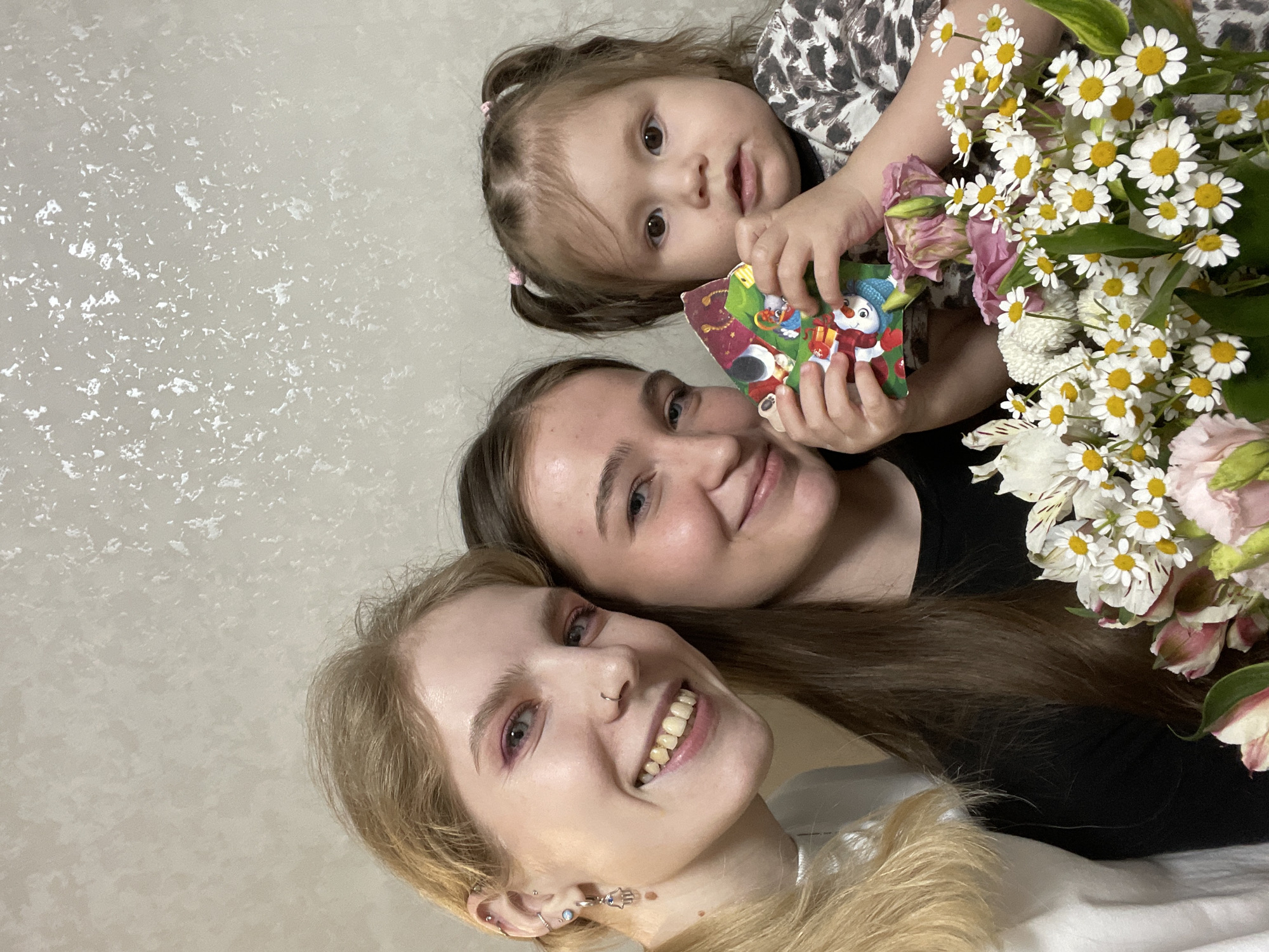 Three people are posing for a photo with a bouquet of flowers. At the top of the image is a young child, holding a small book with cartoon characters on it. Below the child is a young adult woman with dark hair. At the bottom is another young adult woman with blonde hair. The background is a light grey textured wall. There is no indication of the location being Izhevsk, Russia.