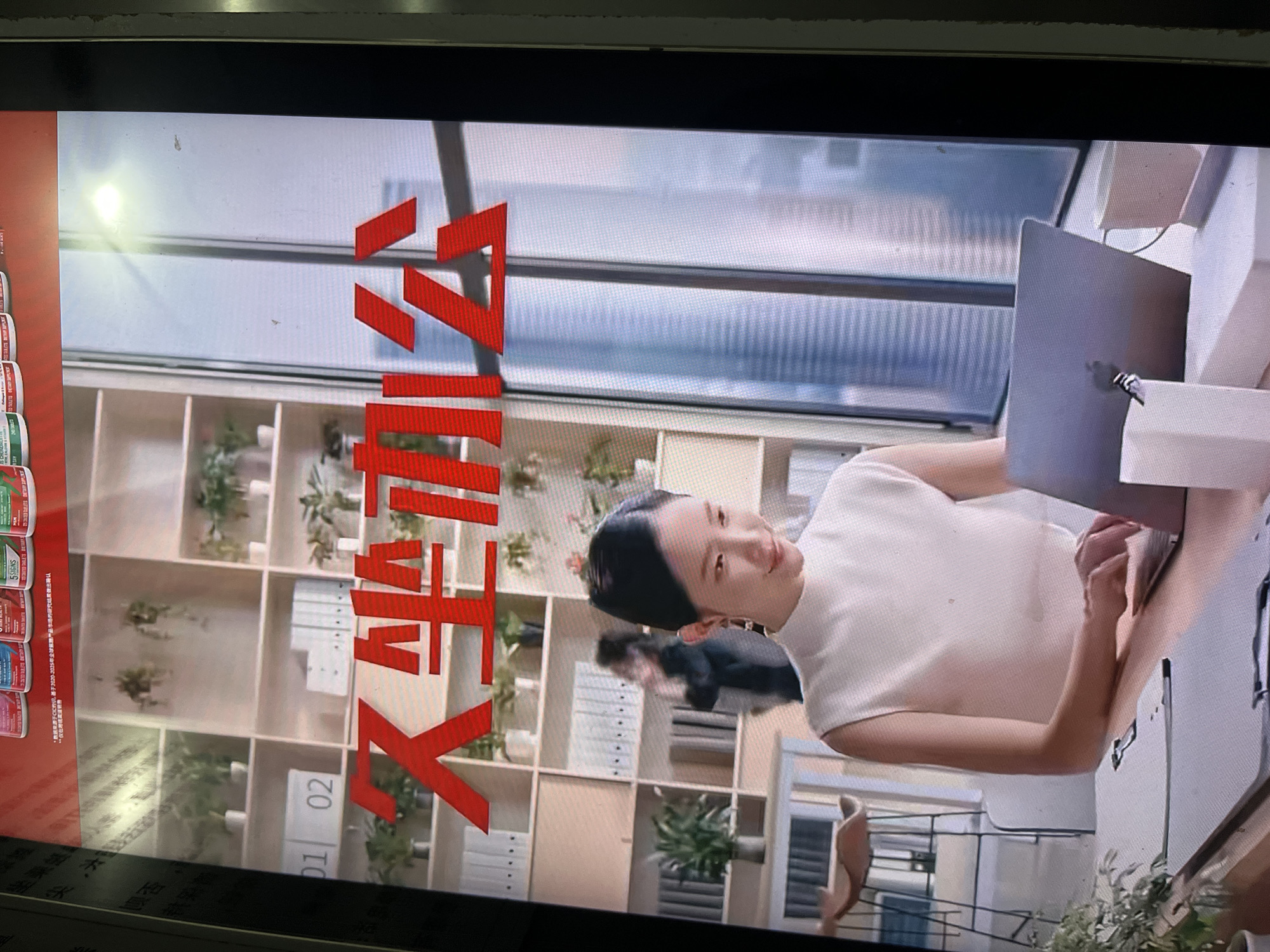 The image is a photograph of a screen, showing a rotated scene of an office or co-working space. The scene is brightly lit by natural light coming through large windows or glass partitions in the background.
The primary subject is a woman with dark hair pulled back, wearing a light-colored, sleeveless top. She is seated at a desk, looking slightly towards the viewer while actively typing on a silver laptop (resembling a MacBook). Her expression is calm and engaged, with a hint of a smile.
In the background, there is a modern modular bookshelf unit filled with potted plants and various files or books. A blurred figure, possibly another person, is visible deeper within the office space. To the far left, partially visible, are several small containers, possibly product packaging, against a red background.
The setting suggests a contemporary and organized work environment. The time of day appears to be daytime due to the bright natural light.
Visible text includes large, stylized red characters prominently displayed in the middle, which the OCR identifies as "义大利" (Yìdàlì, meaning Italy). On the partially visible product containers on the left, the text "5SIGNS" is visible, along with Chinese text: "*数来源于CIC约识,基于2020-2025年全球路产品市场的研究结果做出确认" (meaning "*Data comes from CIC Consultation, confirmed based on research results from the global road product market from 2020-2025."). The numbers "01" and "02" are also visible on the bookshelf compartments.