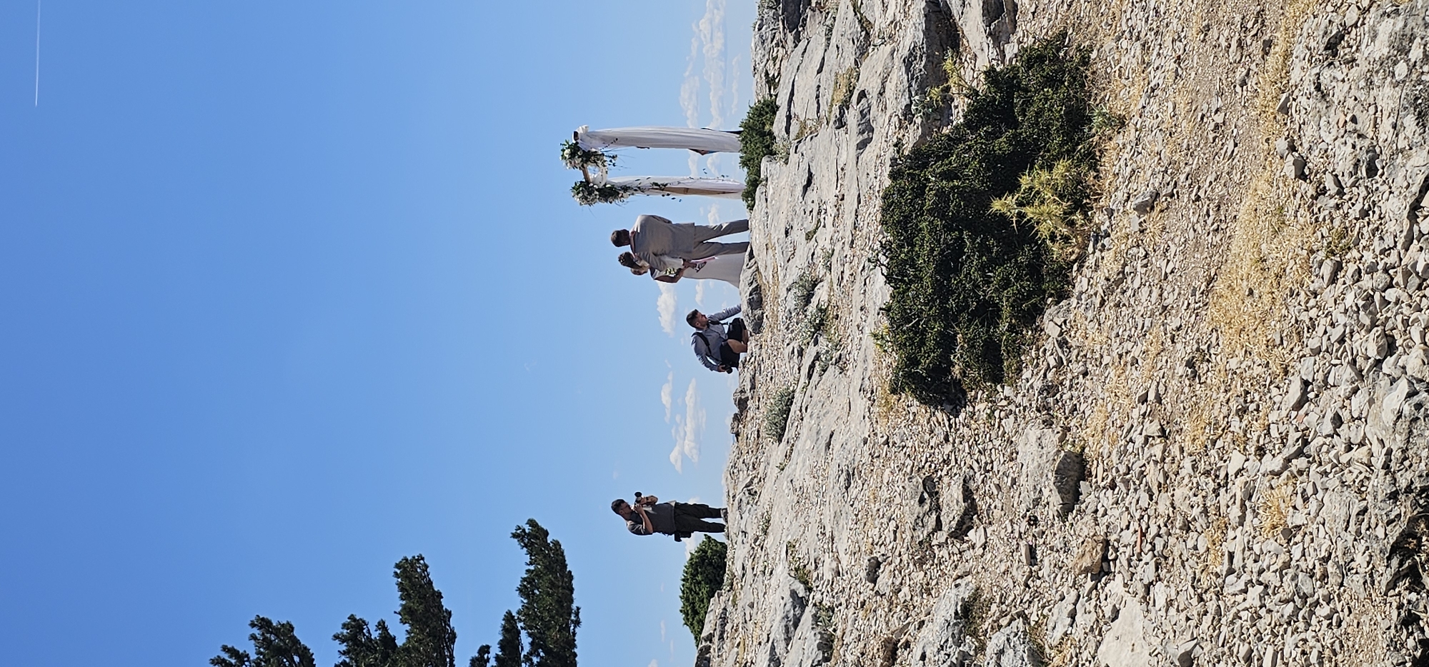 This image captures a wedding ceremony taking place outdoors on a rocky terrain under a clear blue sky with scattered white clouds. The scene is set on a hilltop, likely in the vicinity of Općina Bol, Croatia, given the characteristic rocky landscape and vegetation.
In the center of the frame, a bride in a white wedding dress and a groom in a light-colored suit are standing near a floral archway decorated with white drapery and greenery. The groom is holding a young child.
Two photographers are present, actively documenting the event. One photographer, dressed in a dark shirt and pants, stands to the left of the couple, holding a camera and looking down at it. Another photographer, in a grey shirt and dark shorts, is crouched down in the foreground, closer to the couple, also with a camera.
The environment is dry and arid, with numerous grey rocks and sparse, low-lying green and yellow vegetation. A mature tree with dark green foliage is visible on the far left edge of the image, contributing to the natural, rugged setting. The overall impression is one of a simple, intimate wedding ceremony held in a picturesque, natural landscape.