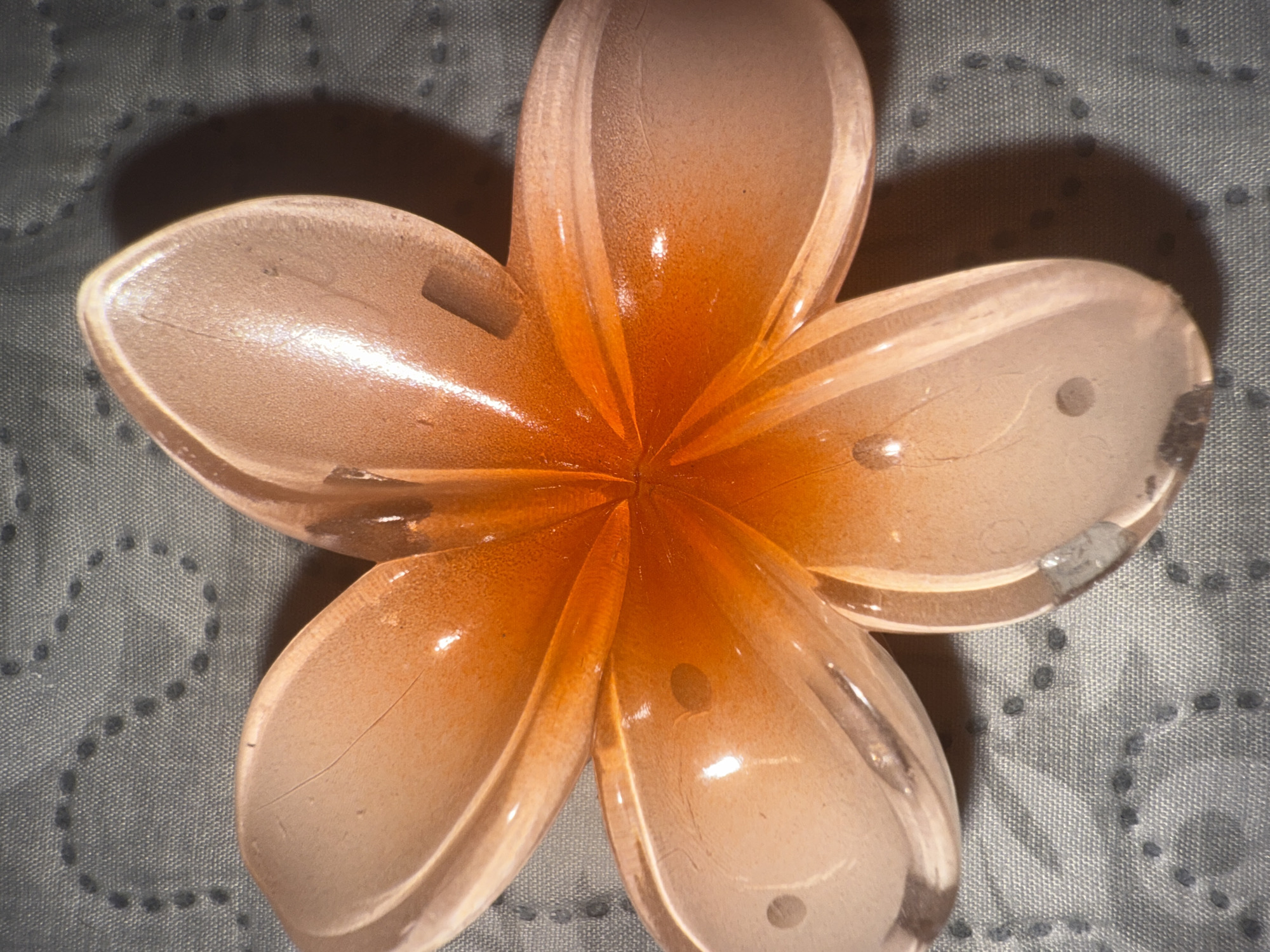 A translucent, five-petaled, flower-shaped object dominates the frame, exhibiting a gradient of peach and orange hues. The object, likely crafted from glass or resin, is brightest at its center, where a vibrant orange radiates outwards, softening to a pale peach at the edges of the petals. The material has a slightly frosted or textured finish, and several petals display fine lines or cracks, along with small, dark inclusions or bubbles. These details are particularly visible in the cropped views.
The flower casts a distinct, dark shadow on the background. The background itself is a light gray fabric with a subtle woven texture. Embellished on the fabric is a repeating pattern of small, dark gray dots, forming curvilinear designs, some resembling partial letters or abstract shapes. The lighting appears artificial, highlighting the object's translucency and creating reflections on its surface. No activity is taking place, and there is no visible text. The setting appears to be indoors, in Varna, Bulgaria.