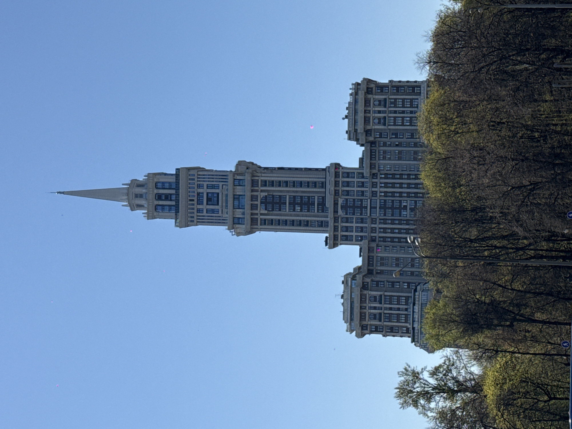 The image shows a tall Stalinist skyscraper against a clear blue sky, situated near trees. It has a central, tapering tower with a spire at the top, flanked by symmetrical, lower wings. The building's facade is light gray, with many windows, and appears to be in Khimki, Russia.