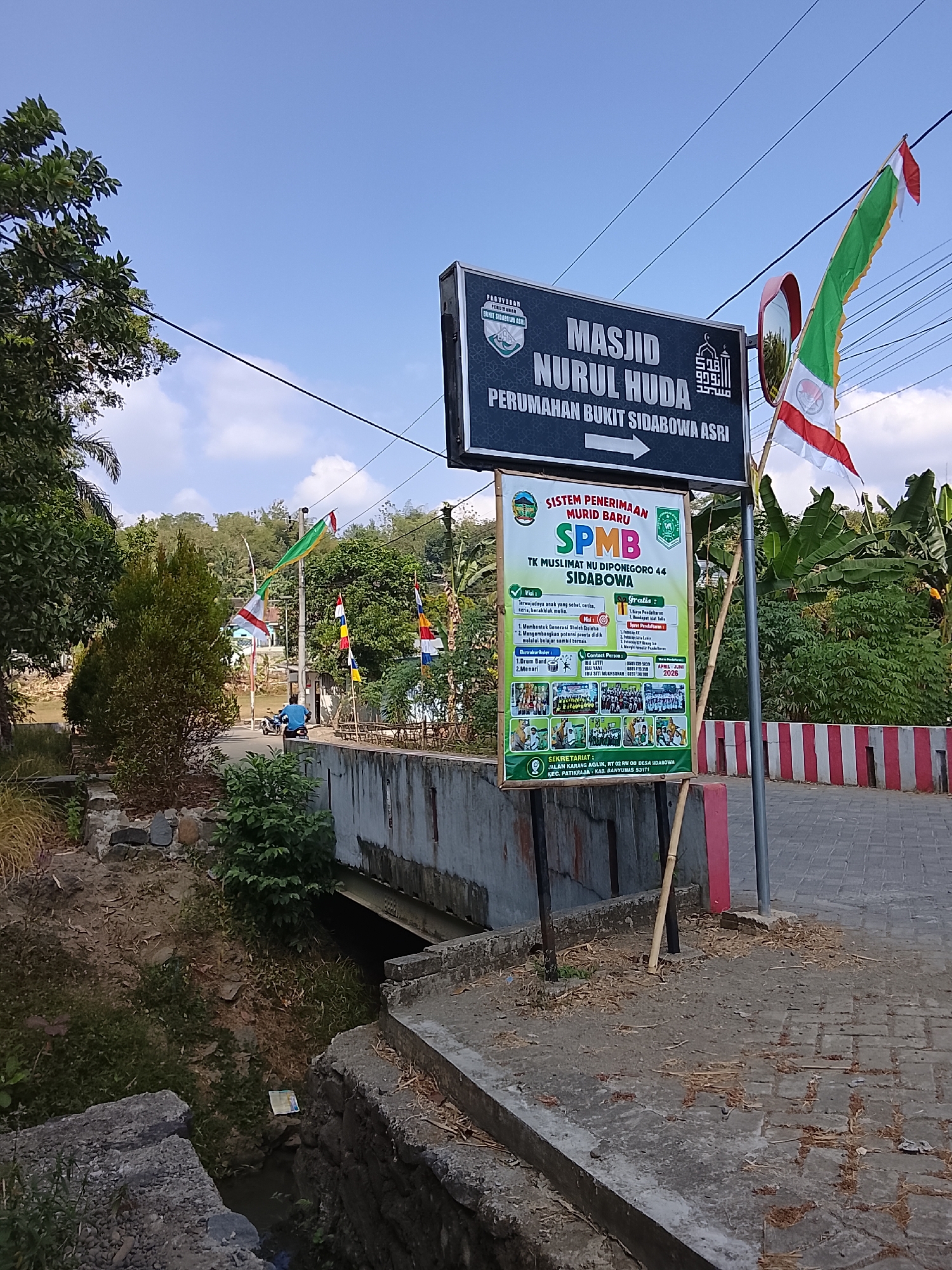 In Sidabowa, Indonesia, a street scene unfolds under a bright, clear blue sky with scattered clouds. A prominent black and dark blue sign announces "MASJID NURUL HUDA" and "PERUMAHAN BUKIT SIDABOWA ASRI," indicating the presence of a mosque and a residential area. Next to this is a smaller, colorful sign detailing "SISTEM PENERIMAAN MURID BARU SPMB TK MUSLIMAT NU DIPONEGORO 44 SIDABOWA," which translates to "New Student Admission System SPMB Muslimat NU Kindergarten Diponegoro 44 Sidabowa." This sign includes information on school fees, free services, extracurricular activities like drum band and dancing, contact information, and the school's address at Jalan Karang AGLIK, RT 02 RW 08 Desa Sidabowa, Patikraja, Kabupaten Banyumas 53171.

Several flags are visible: a large red and white Indonesian flag with a green trim attached to a pole, and smaller colorful flags—yellow, white, blue, and red—along the side of the road. A person on a blue motorcycle is traveling down the paved street. The environment features lush green trees, a concrete bridge over a small waterway, and textured paving stones on the roadside. The overall scene depicts a typical day in a residential and community area in Indonesia.