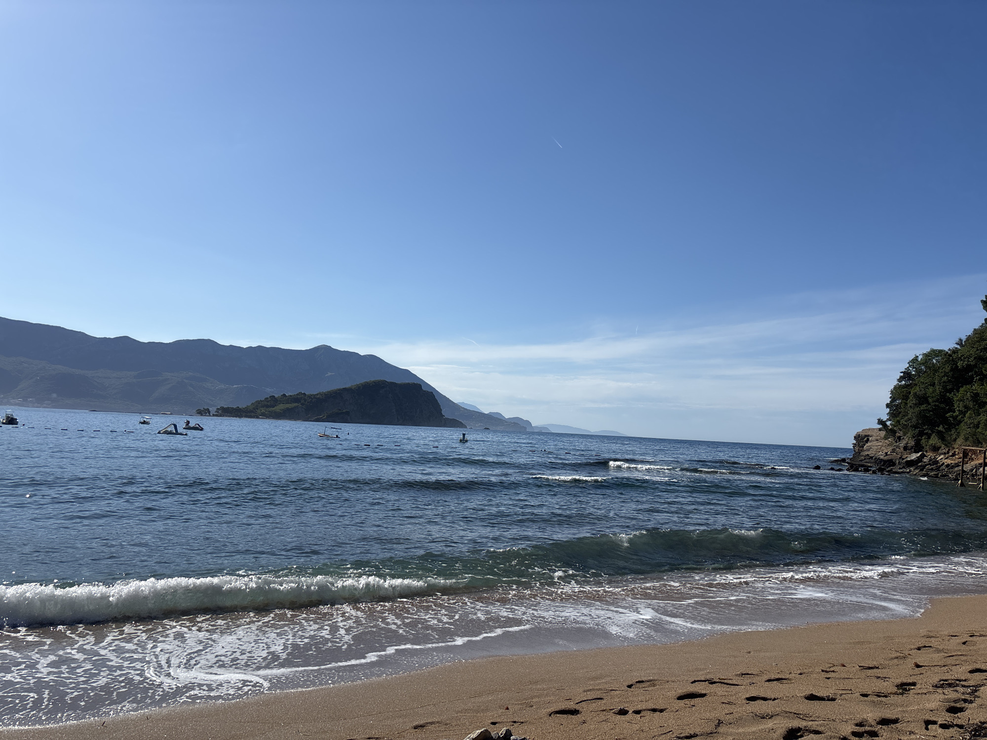 The image depicts a tranquil beach scene with a clear blue sky and calm sea. In the foreground, a sandy beach is visible with gentle waves lapping at the shore, leaving trails of white foam. Several footprints are imprinted on the sand. In the middle ground, the blue sea stretches out, with a few small boats and what appear to be inflatable watercraft scattered across the water. A line of buoys can be seen in the distance. The background features a range of lush green mountains, with one prominent island-like landmass jutting out into the sea. To the right of the frame, a rocky cliff covered in dense green foliage overlooks the water, with some wooden structures visible near the shoreline. The overall atmosphere is peaceful and idyllic, suggesting a pleasant day at the beach.