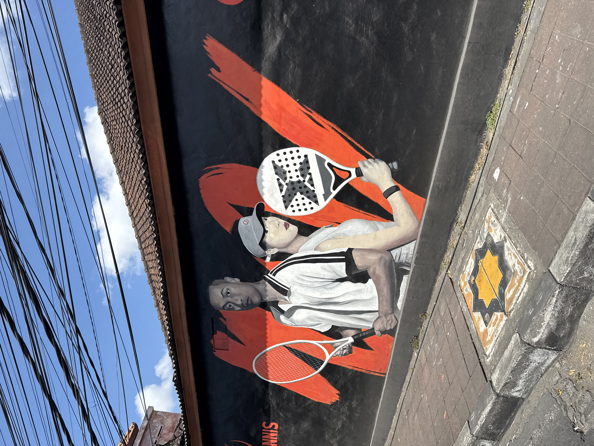 On a sunny day in North Kuta, Indonesia, a mural is painted on a black wall. The mural depicts two individuals, a man and a woman, who appear to be athletes. The man, on the left, wears a white and black tennis outfit and holds a tennis racket. The woman, on the right, is seen from the shoulder up, wearing a grey top and a grey cap, and holding a paddle. Red paint strokes, resembling brush strokes or abstract shapes, are visible on the wall behind the figures. The mural extends across the wall, with the word "TENNIS" visible on the left side. The wall is situated above a sidewalk made of grey concrete and red bricks, with a decorative yellow and black tiled square set into the pavement. The street in front of the sidewalk is paved with asphalt. Overhead, a dense network of black electrical wires crisscrosses the bright blue sky, interspersed with scattered white clouds. The tiled roof of a building is visible above the mural.