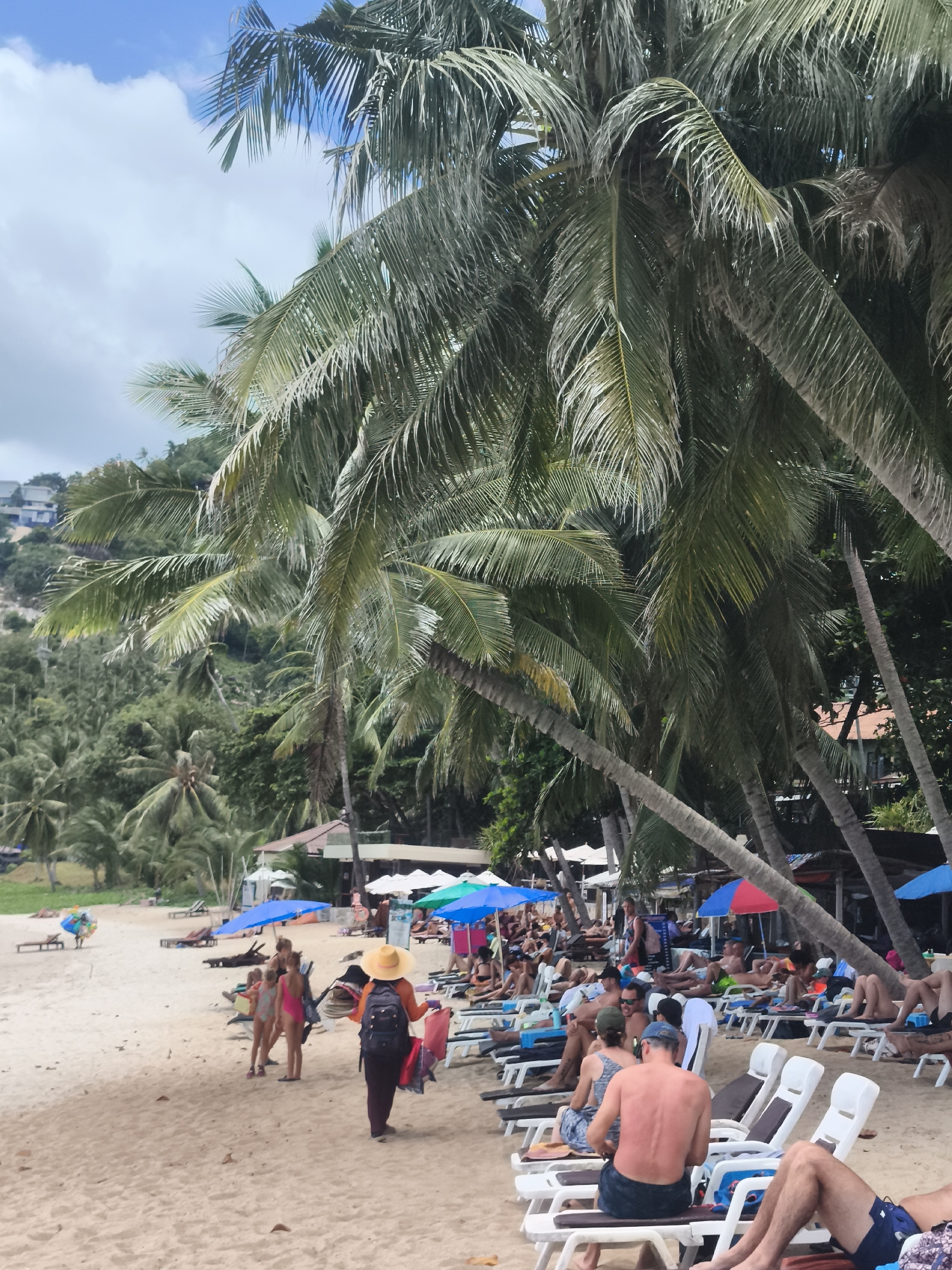 The image shows a busy beach scene in the city of Ko Samui District, Thailand, on a cloudy day with palm trees dominating the foreground. People are relaxing on lounge chairs and walking on the sand. In the background, there are buildings and lush green hills. A vendor wearing a straw hat and carrying bags is walking towards the camera. Other people in the image are wearing swimsuits and casual beachwear. Several colorful umbrellas provide shade.
