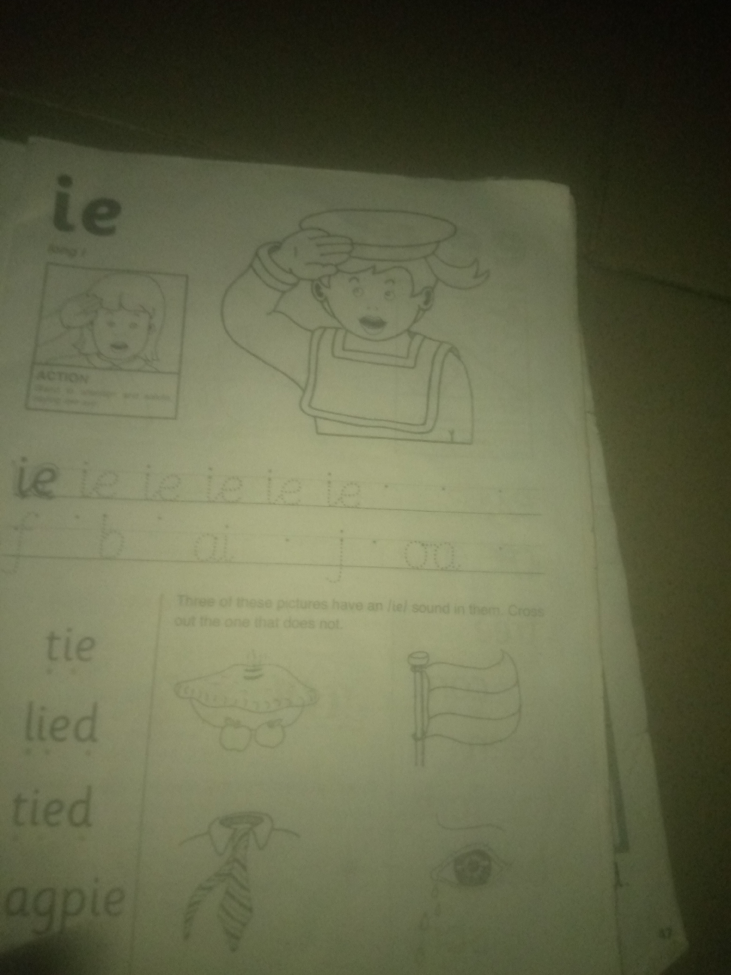 The image shows a page from a workbook, likely for young children learning phonics. The page is predominantly white with some dark smudges and shadows, suggesting poor lighting or image quality.
At the top left, the letters "ie" are prominently displayed in a large, bold font. Below this, a small box contains a line drawing of a child's face with an open mouth and wide eyes, labeled "ACTION." To the right of this, a larger drawing depicts a child wearing a sailor's hat and uniform, with one hand raised to their forehead as if saluting or shielding their eyes.
Below these illustrations, the letters "ie" are repeated multiple times in a smaller, cursive font, suggesting practice writing or recognition. Further down, lines are drawn with dotted letters "f", "b", "ai", and "j" for tracing practice.
To the left of the page, there are several words written in a clear, sans-serif font: "tie," "lied," "tied," and "agpie."
The bottom portion of the page contains instructions: "Three of these pictures have an /ie/ sound in them. Cross out the one that does not." Below this text are three line drawings:
1. A simplified drawing that appears to be a hat with apples hanging from it.
2. A drawing of a flag on a pole.
3. A drawing of a tie.
4. A drawing of an eye with a tear falling.
The page is slightly creased and wrinkled in places. A faint watermark or stamp is visible on the right edge, showing the number "47".
There is no information in the image that relates to the city of Jauro Yinu, Nigeria. The description is limited to the visual content of the provided image.