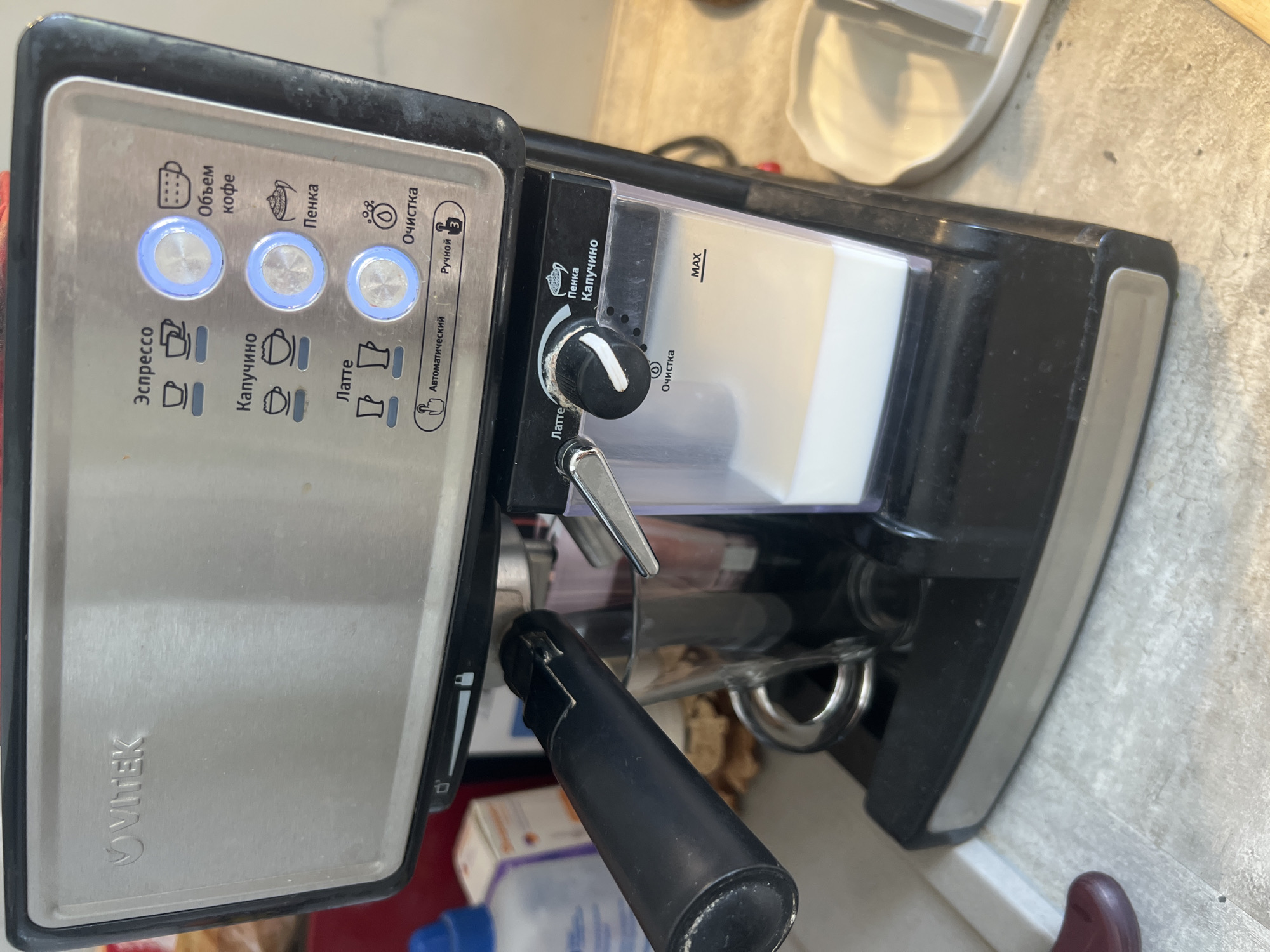The image shows a close-up of a VITEK brand espresso machine on a kitchen counter. The machine has several buttons and indicators, including options for "Espresso," "Cappuccino," and "Latte." There is a milk frother attachment visible to the right of the machine, with a dial for adjusting settings and a "MAX" fill line indicated on the milk container. A glass cup is positioned beneath the coffee dispenser, with a dark liquid inside, possibly coffee or tea. The counter surface appears to be tiled and speckled. In the background, a blue bottle and a box of what looks like medication or cleaning supplies are partially visible. No people are present in the image. The scene is indoors and appears to be a domestic setting. The location of Volgograd, Russia is not identifiable from the image.