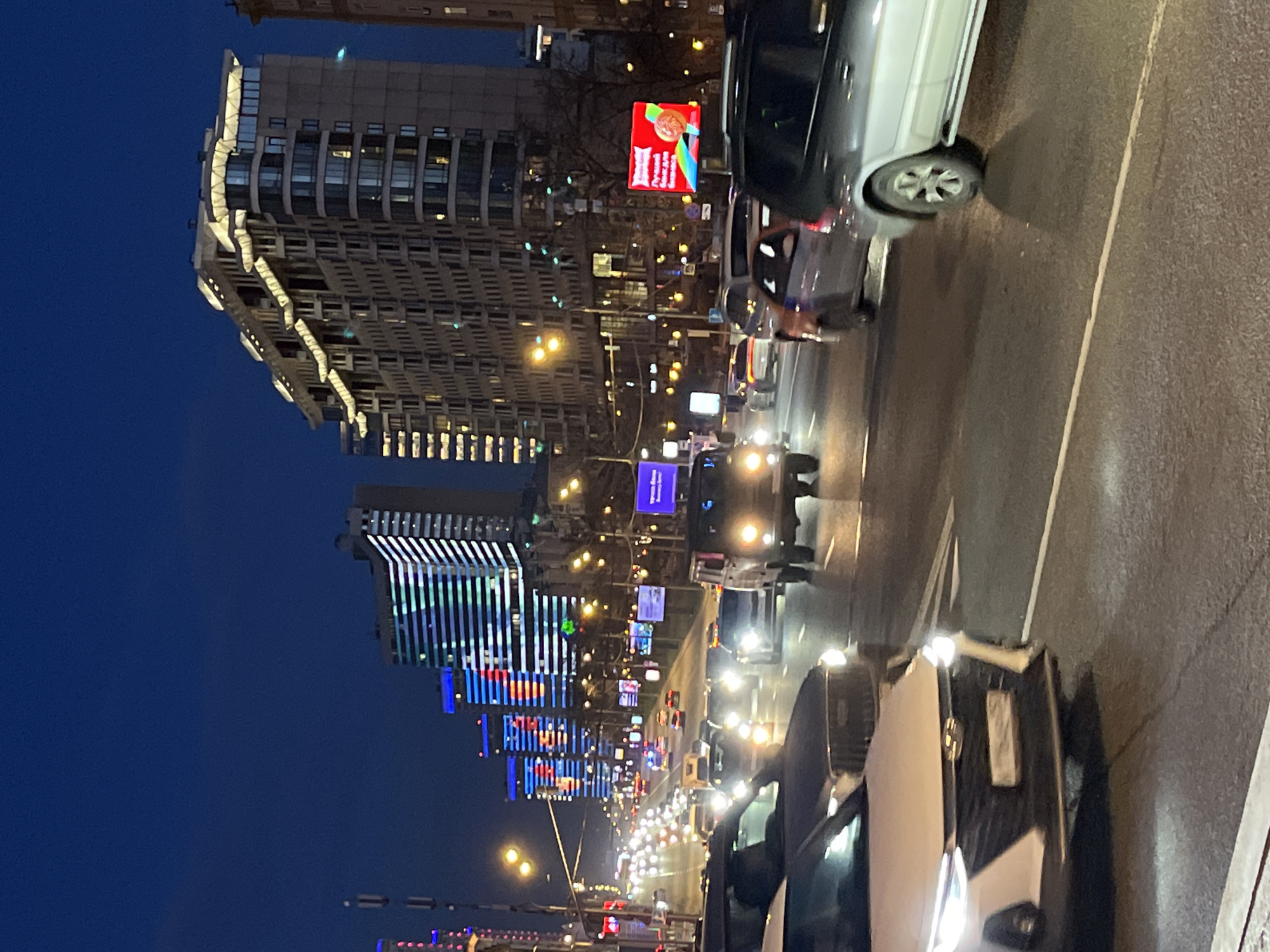 The image shows a street scene in Moscow, Russia at night. The street is lined with tall buildings, some of which are illuminated with bright neon lights. Traffic is moderate, with cars and trucks visible on the road. The sky is dark blue. There are no people visible. A large advertising screen is visible on one of the buildings.