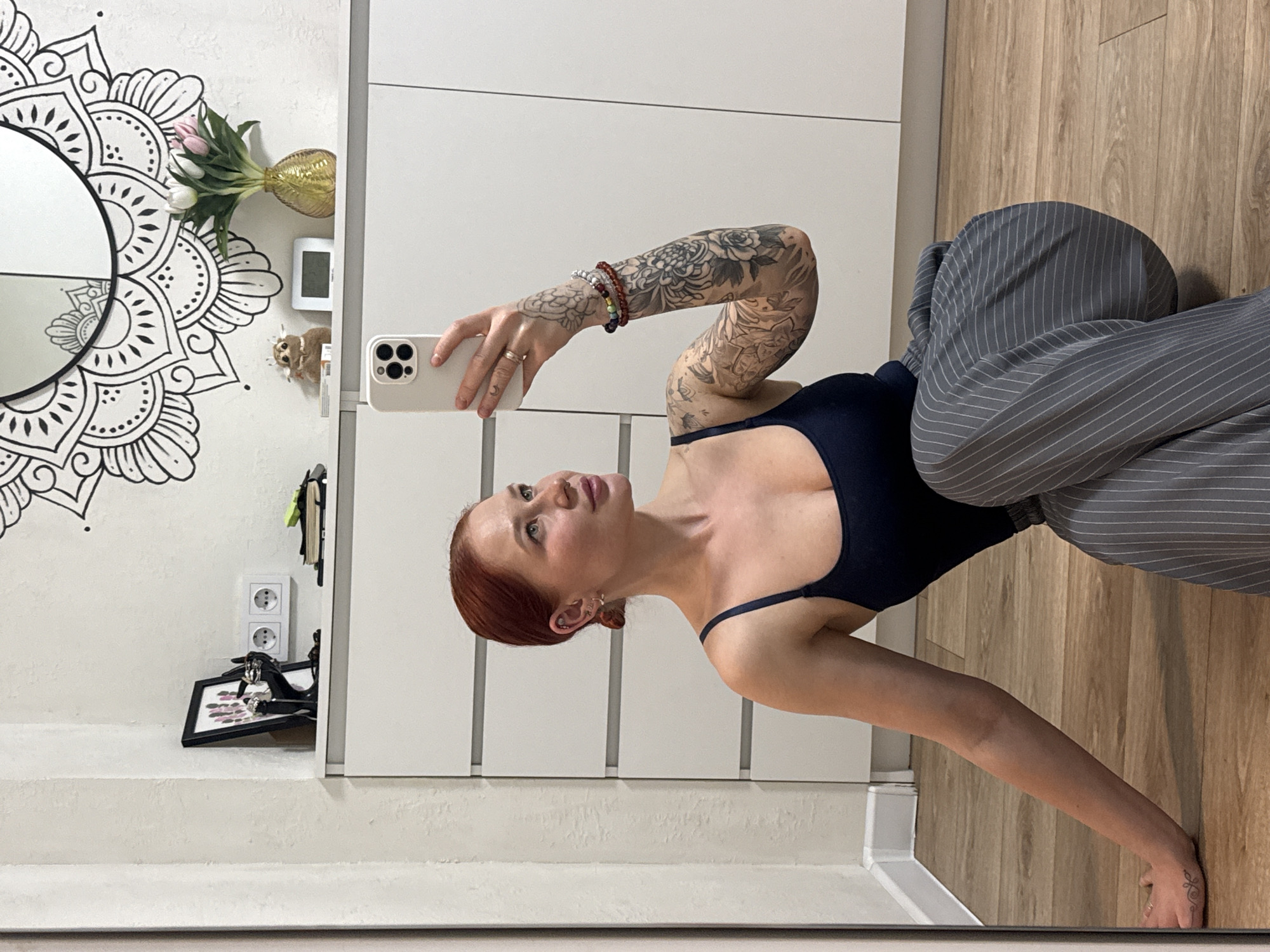 A red-haired woman is taking a selfie in a mirror. She is wearing a dark blue tank top and gray pinstripe pants. Her left arm is raised, holding a white smartphone. Tattoos are visible on her left arm.
The background includes a white wall with a mandala-style design, a cabinet or wardrobe, a wooden floor, a vase of tulips, a small digital clock, a picture frame on the floor, and a double electrical outlet. The reflection in the mirror suggests the image was taken indoors.
It is not possible to definitively confirm the location in the city of Rostov-on-Don, Russia, based solely on the visual information provided.