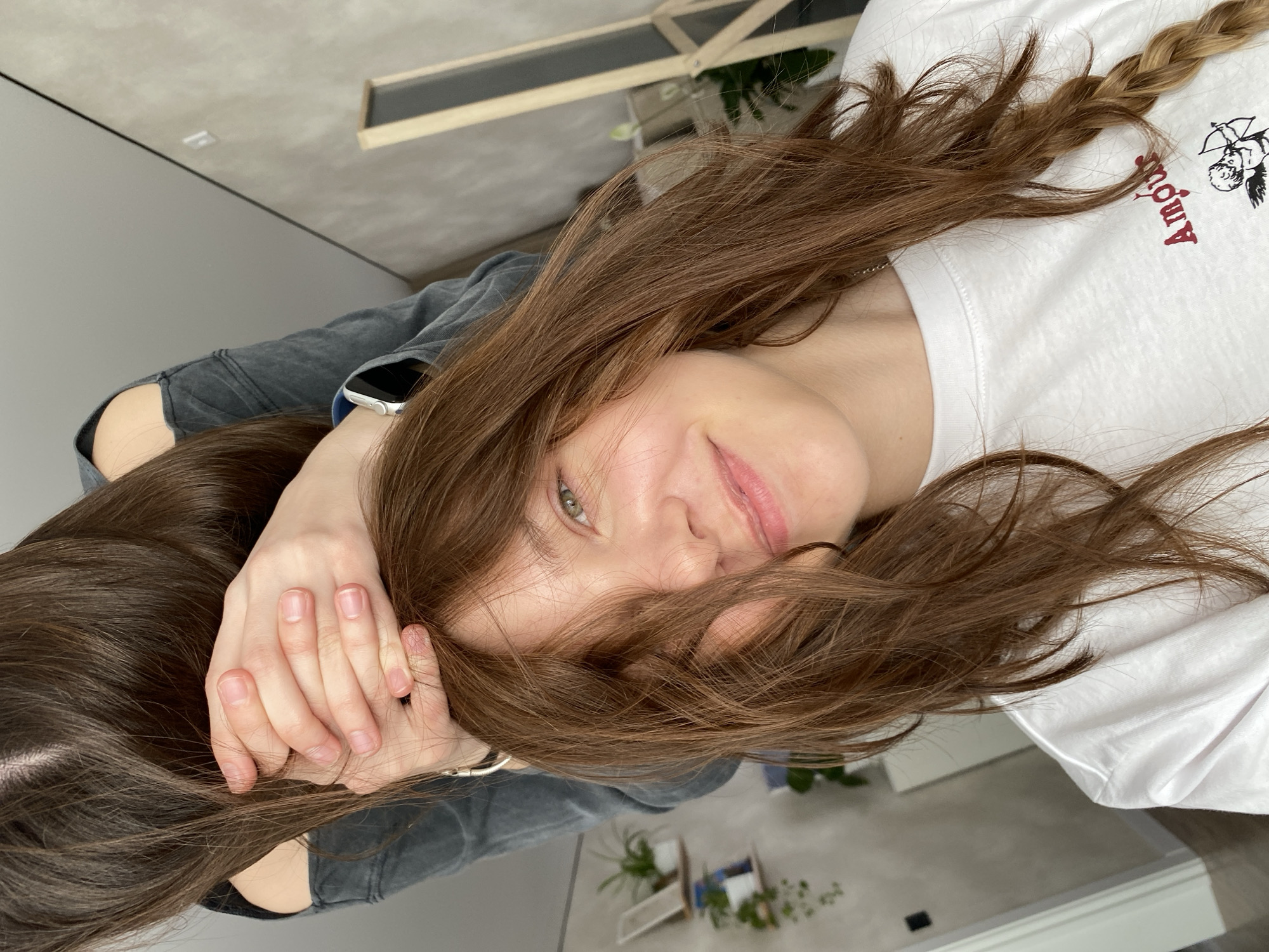 A young woman with long, light brown hair poses for a close-up, eye-level shot. Part of her hair is styled in a braid. She wears a white t-shirt with the word "Amour" printed on it, as well as a grey jacket. Her hands are raised near her face. The background includes what appears to be a room with neutral tones, a window, and potted plants. The location is unknown but is assumed to be Votkinsk, Russia.