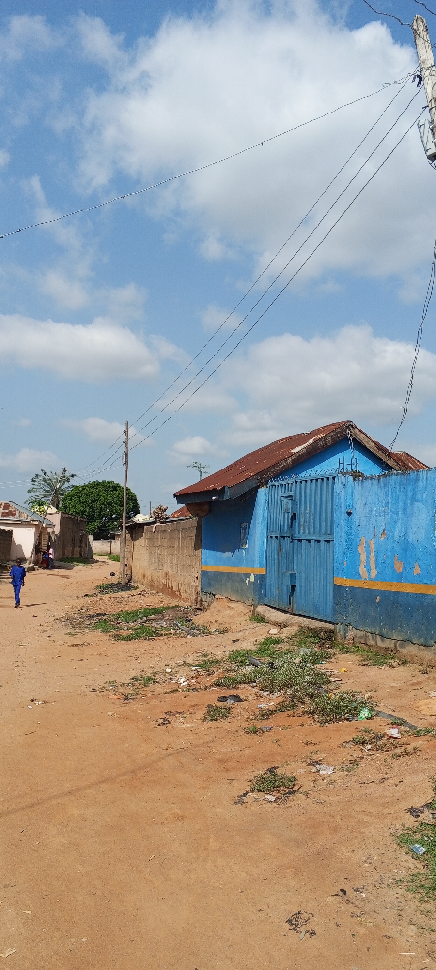 The image shows a dirt road in Jalingo, Nigeria, on a bright, cloudy day. The foreground is dominated by the sandy, reddish-brown road, scattered with debris and patches of sparse green vegetation. In the mid-ground, a person in a blue uniform walks away from the camera down the road. To the right, a blue building with a rusty corrugated metal roof stands behind a low, rough concrete wall. The building has a large, blue metal gate and a yellow stripe running along its lower section. To the left of the building, further down the road, are more buildings, some with light-colored walls and corrugated roofs, and a large, leafy green tree. Several utility poles with power lines crisscross the sky. The overall scene depicts a typical street in a rural or semi-urban area in Nigeria, with a lived-in, somewhat unkempt appearance. The bright sky and clear visibility suggest it is daytime, likely late morning or early afternoon. There is no visible text in the image.