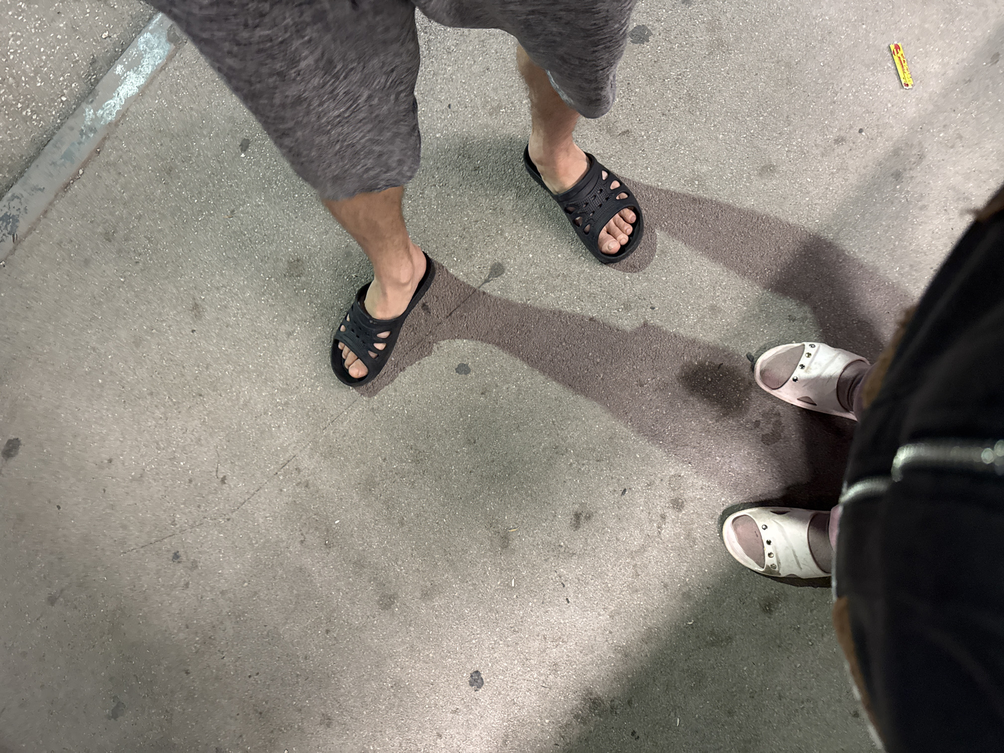 The media file shows a top-down view of four feet wearing sandals on a textured concrete surface. Two individuals are visible, one wearing dark grey shorts and black slide sandals, and another wearing black pants and white slide sandals. The person in shorts is standing with their left foot slightly forward and their right foot behind. The person in black pants is positioned further to the right, with their feet close together. The ground appears to be outdoors, possibly a sidewalk or paved area, given the texture and the presence of a metal edge on the left side of the image. There is a faint shadow cast on the ground, suggesting it is daytime. A small yellow object with red markings is visible on the ground in the upper right quadrant of the image. The overall lighting is somewhat dim, but the scene is clearly illuminated. No specific emotions are conveyed, and the activity appears to be casual, with the individuals standing. The location is specified as Добровское сельское поселение, Ukraine.