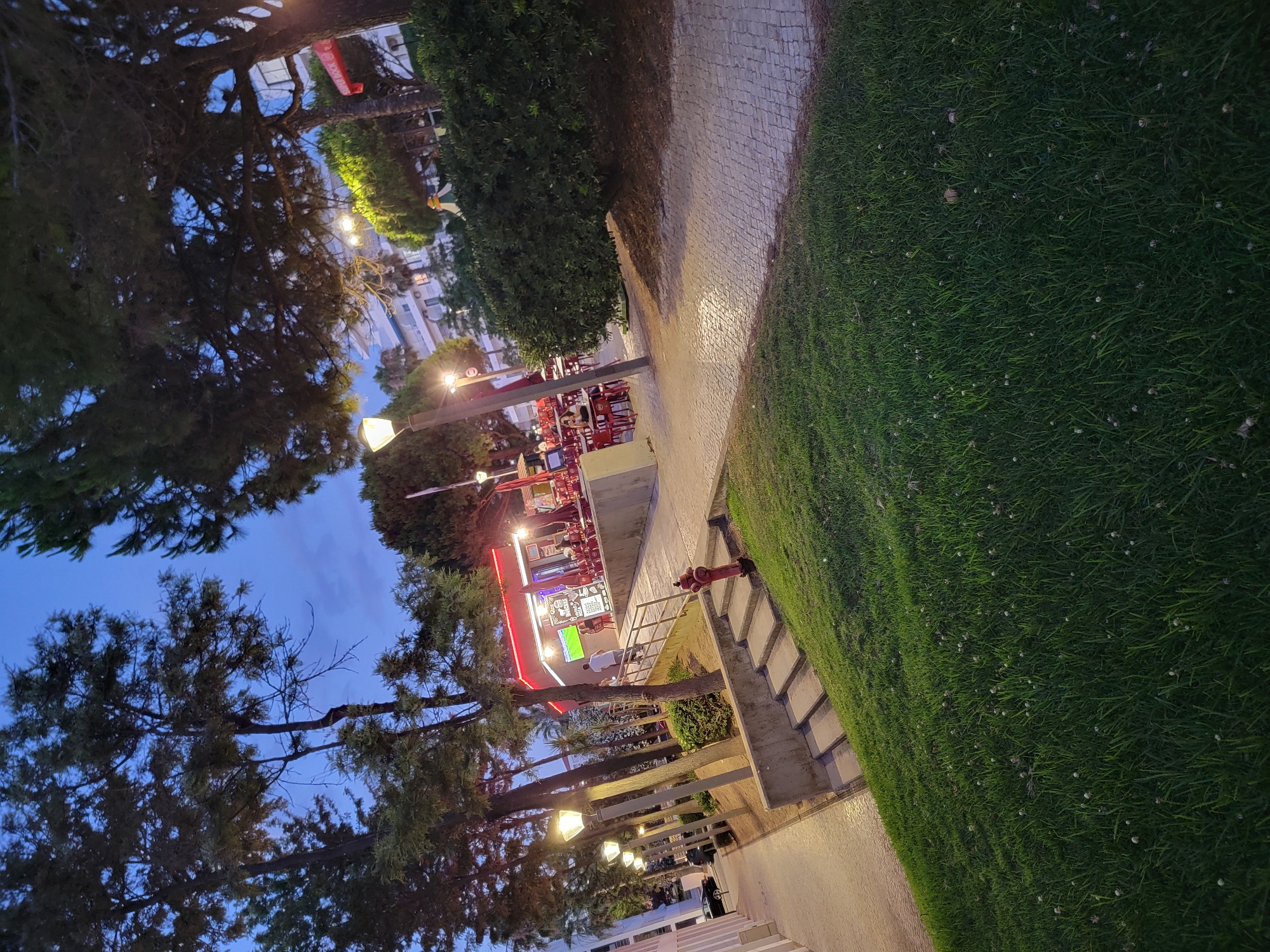 The image depicts an outdoor scene at dusk in Quarteira, Portugal, likely an area with commercial establishments and green spaces. In the background, a building with several floors is visible, illuminated by artificial lights. In the mid-ground, there is a small building with a red neon sign and an outdoor seating area with red tables and chairs, suggesting a restaurant or cafe. A few people are present, including individuals standing and seated at the tables. Adjacent to the seating area is a large, manicured hedge. A paved walkway with cobblestone patterns runs through the scene, lined with lampposts. To the left of the walkway, a grassy slope is visible, dotted with small white flowers. A red fire hydrant is positioned on a concrete structure with stairs leading up to the walkway. A dark-colored car is parked on the street to the left. Trees with dark green foliage are prominent throughout the image, with their branches casting shadows. The sky above is a deep blue, indicating twilight.